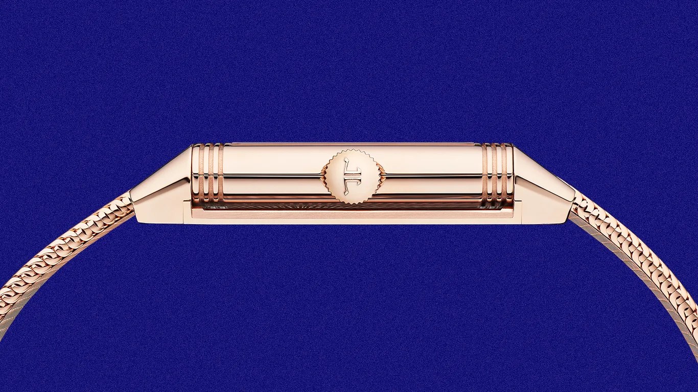 Rose gold watch crown with a textured edge and a logo on a matching rose gold mesh strap against a blue background.