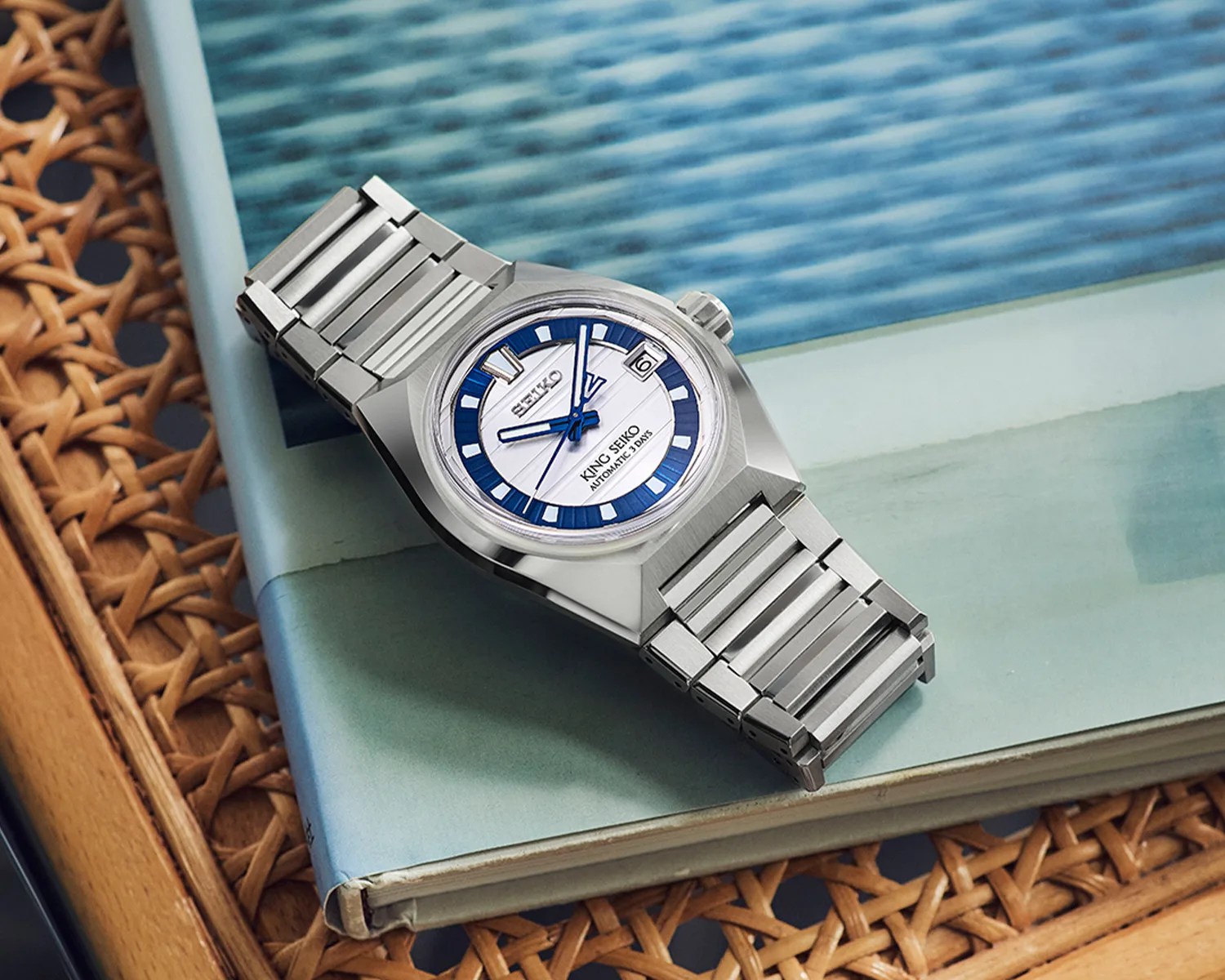 Silver stainless steel King Seiko wristwatch with blue and white dial resting on a book with a woven surface underneath.