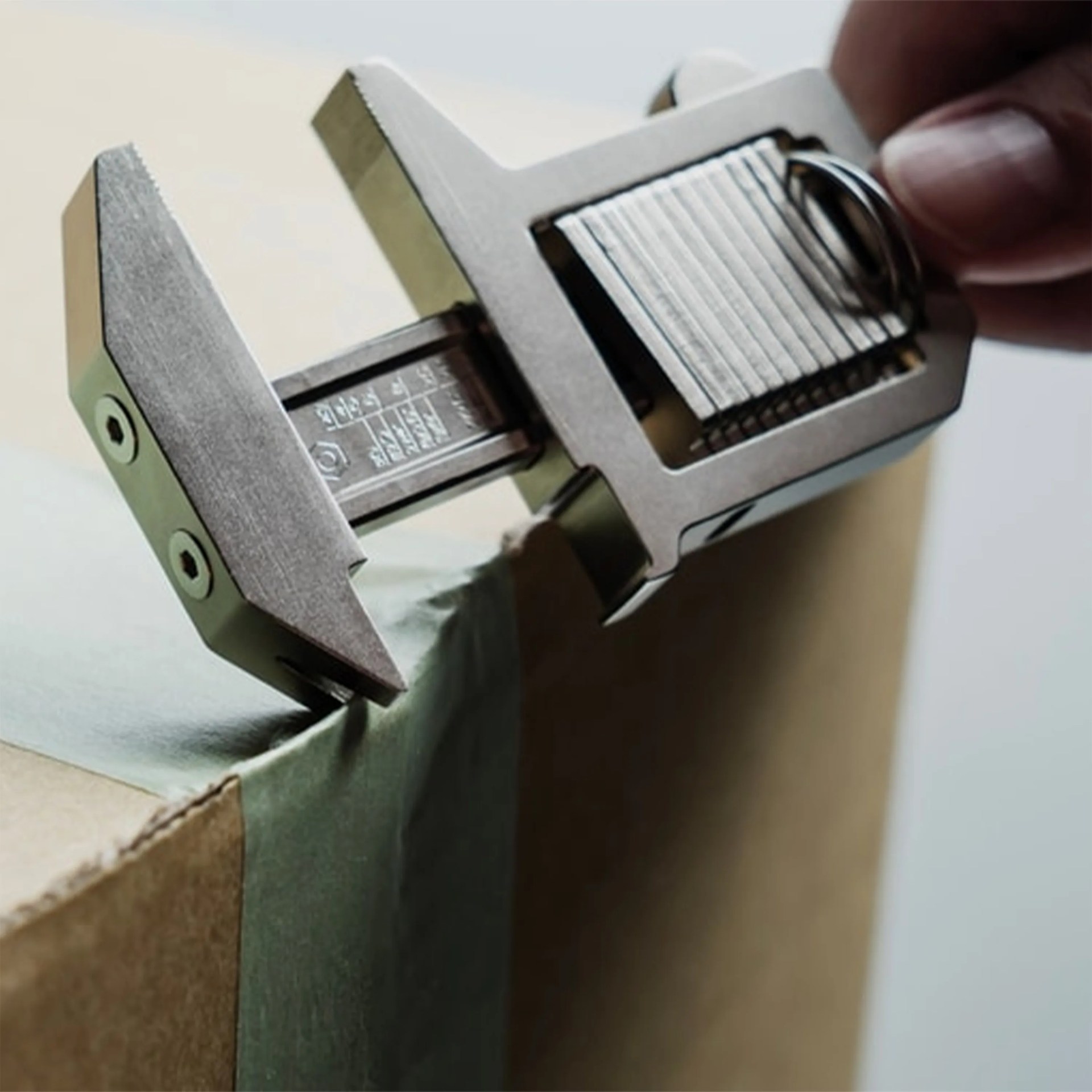 Close-up of a metal caliper measuring the thickness of cardboard with green tape on the edge.