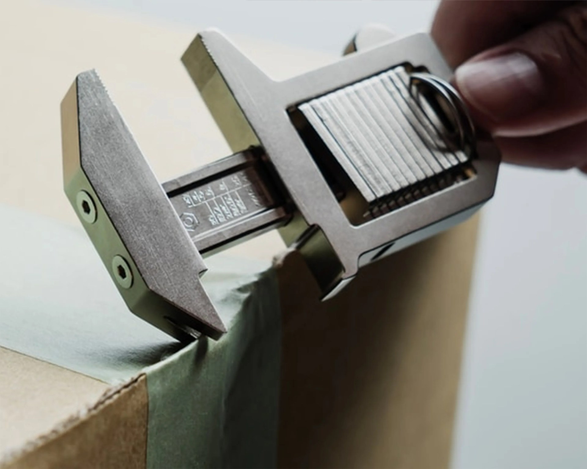 Close-up of a hand adjusting a metal caliper measuring the thickness of green tape on a cardboard box edge.