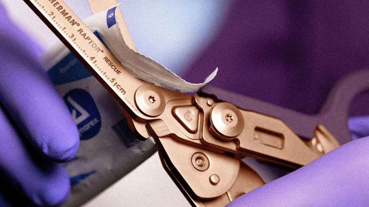 Close-up of a gold-colored Leatherman Raptor Rescue tool cutting open a package, held by hands in purple gloves.