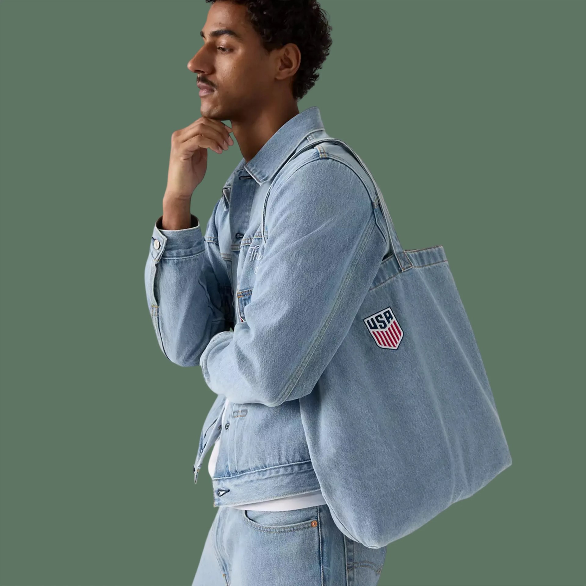 Man wearing light blue denim jacket and jeans with a matching denim tote bag featuring a USA patch.