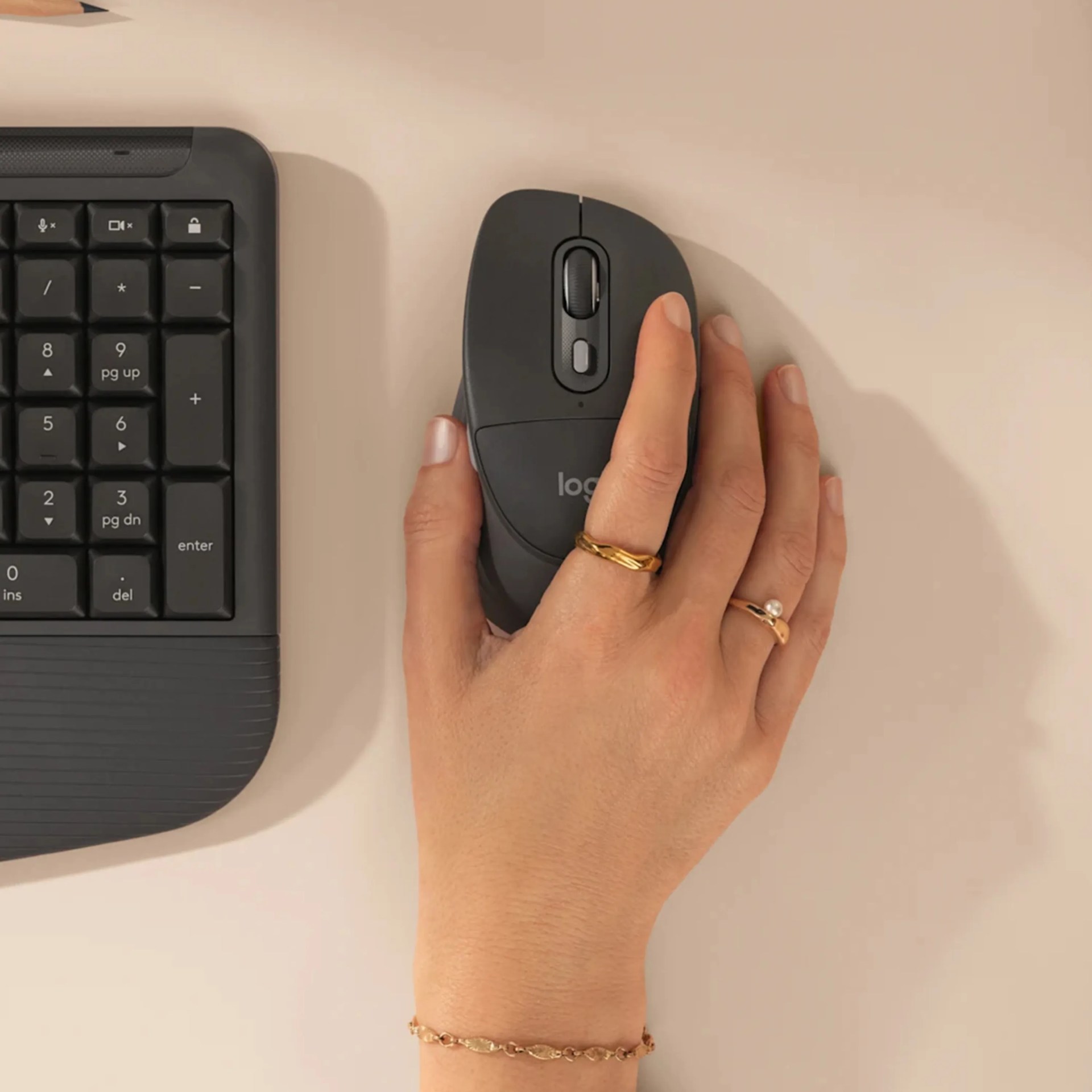 Hand with gold rings and bracelet using a black Logitech wireless mouse next to a black keyboard on a beige surface.