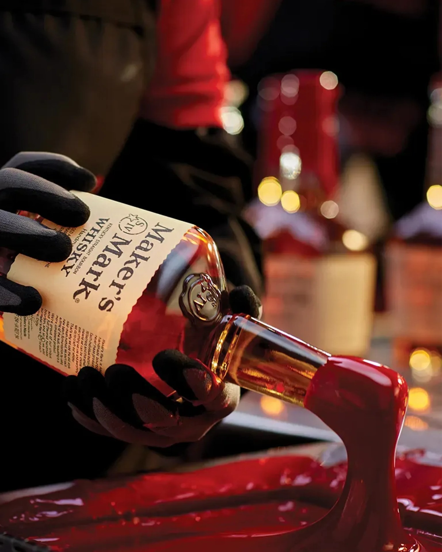 Hands wearing black gloves holding a Maker's Mark whiskey bottle with red wax dripping from the top.