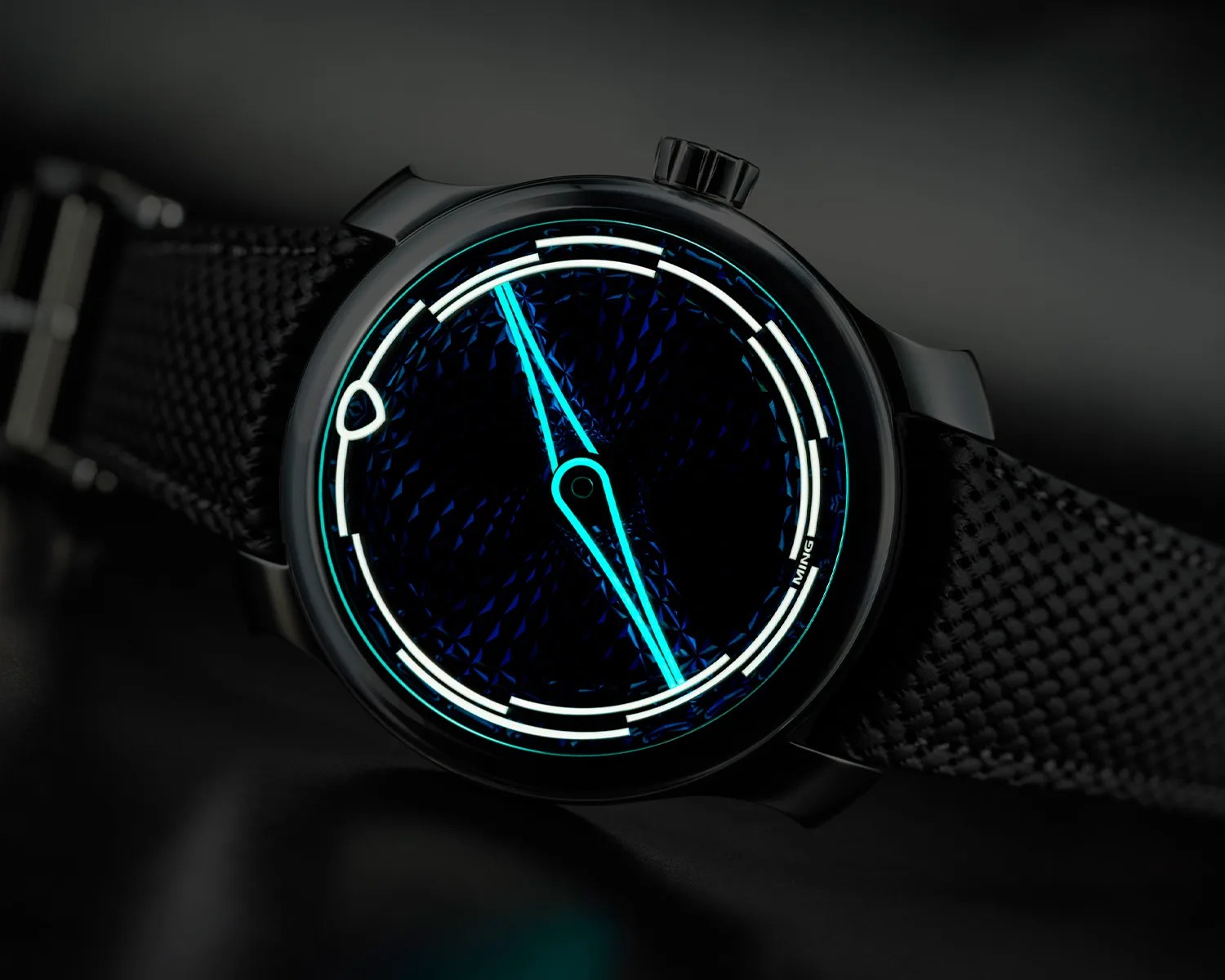 Black wristwatch with glowing blue hands and white hour markers on a textured dark dial and black woven strap.
