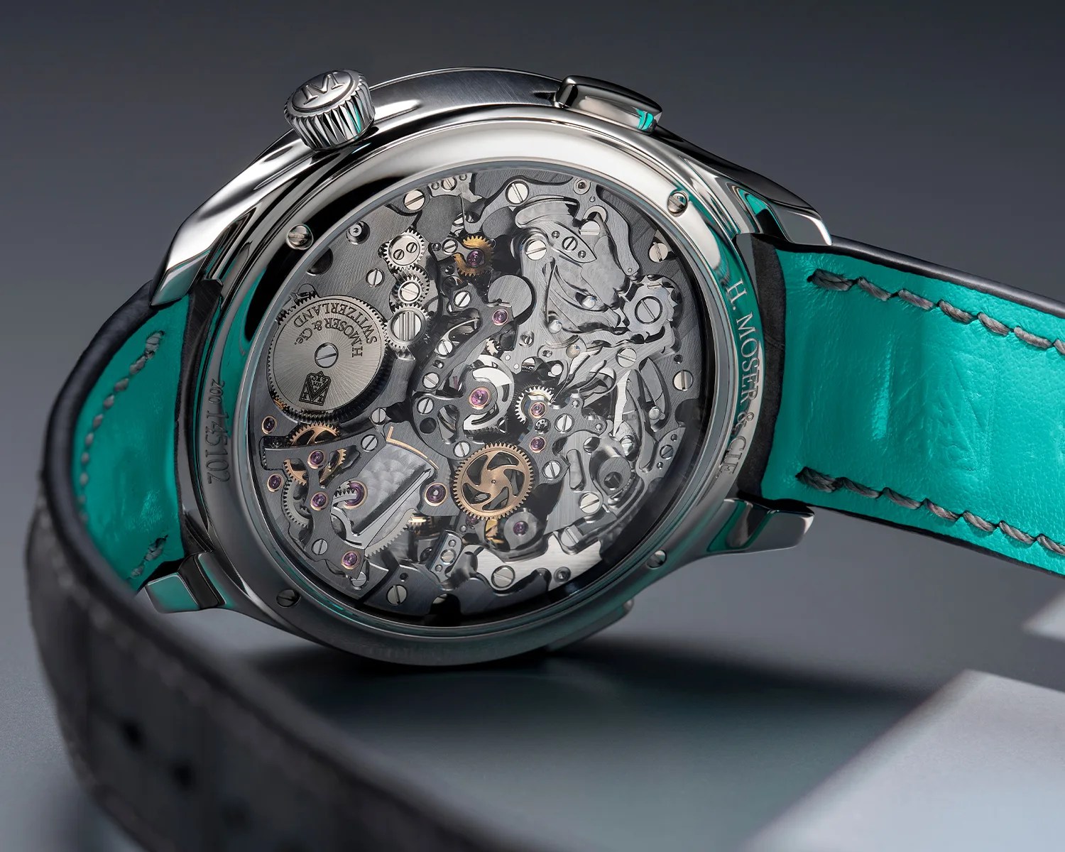 Close-up of a silver H. Moser & Cie watch back showing intricate mechanical gears and a turquoise leather strap.
