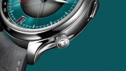 Close-up of a silver watch crown with an engraved "M" on a teal dial and gray leather strap.