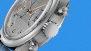 Close-up of a silver chronograph watch with blue and gold hands and a blue leather strap against a blue background.