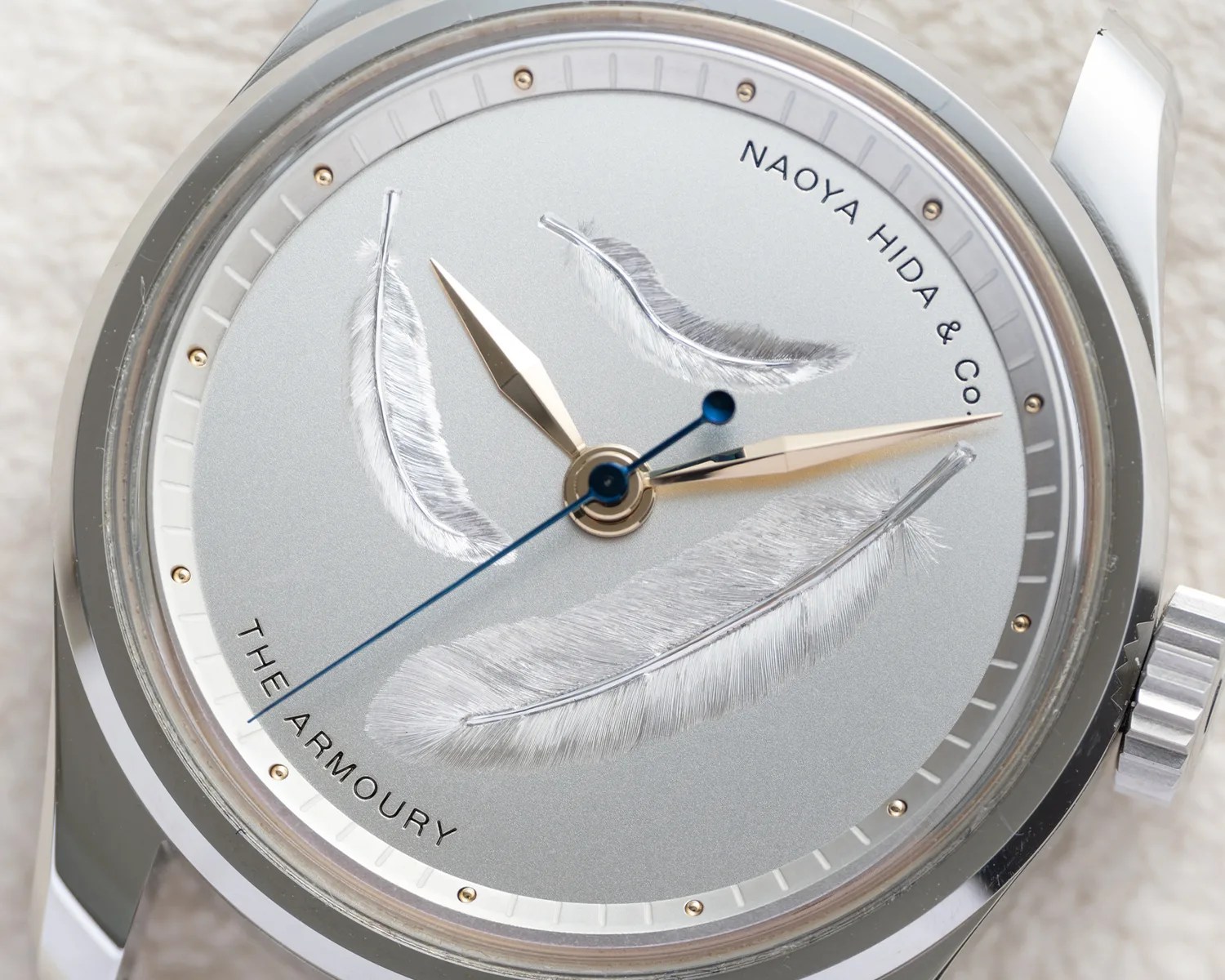 Silver wristwatch dial with engraved feathers, gold hour and minute hands, and a blue second hand.