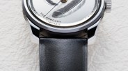 Close-up of a silver watch face with a feather design and black leather strap on a textured white surface.
