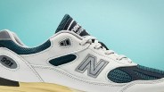 White and teal New Balance 991 sneaker with mesh panels and beige sole against a light blue background.