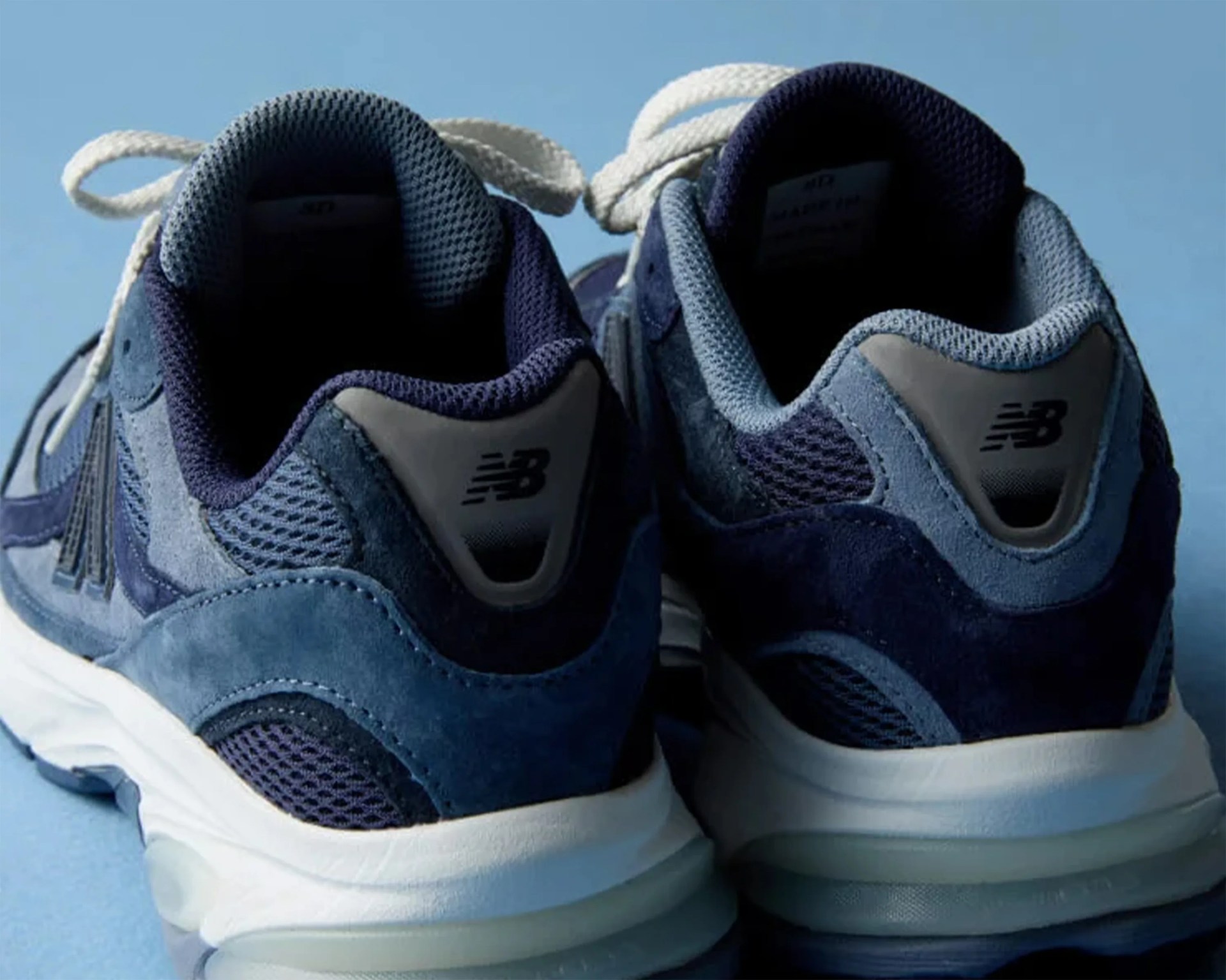 Pair of blue New Balance sneakers with white soles viewed from the back on a blue surface.