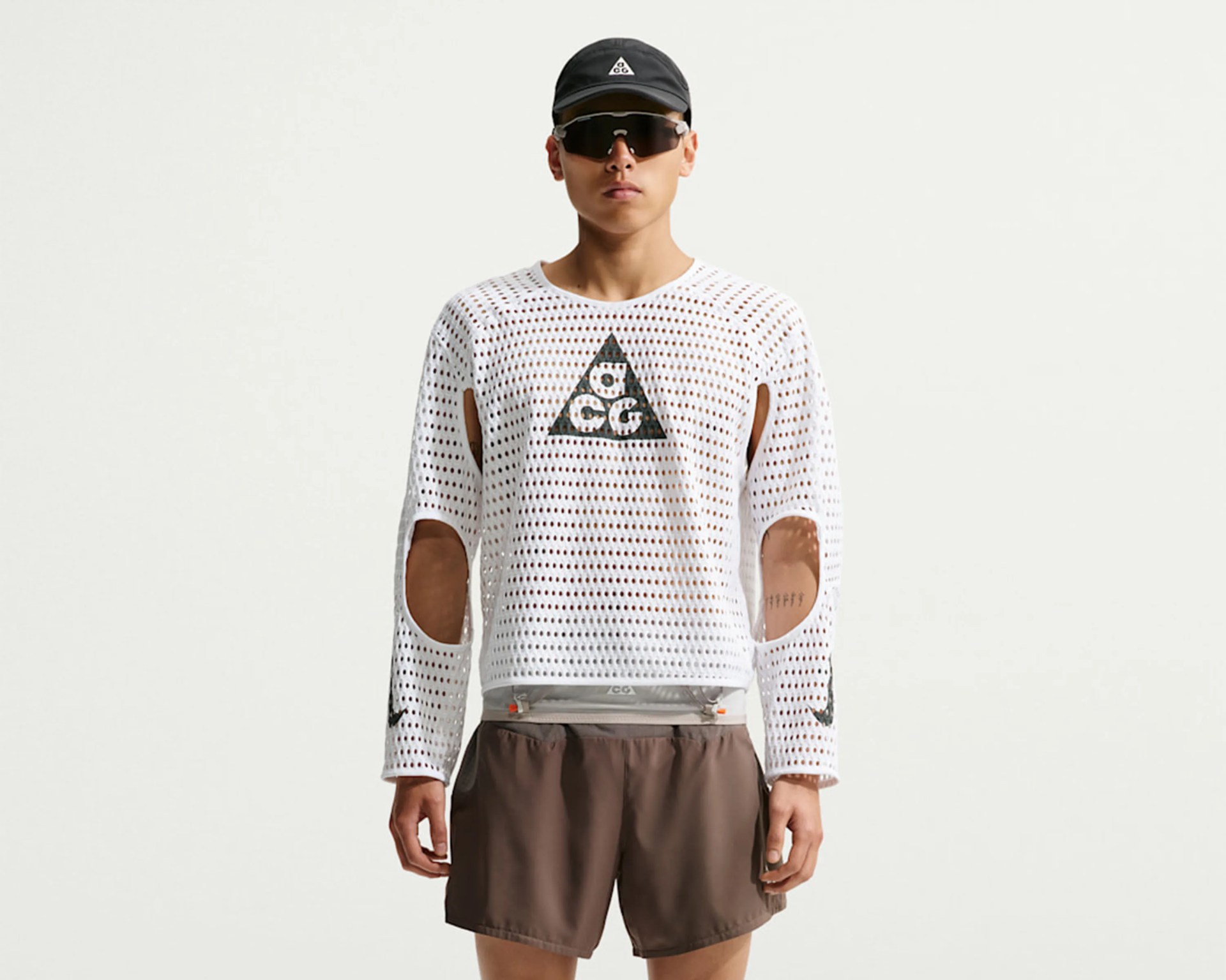 White long-sleeve mesh shirt with ACG logo and elbow cutouts worn with brown shorts and black cap.