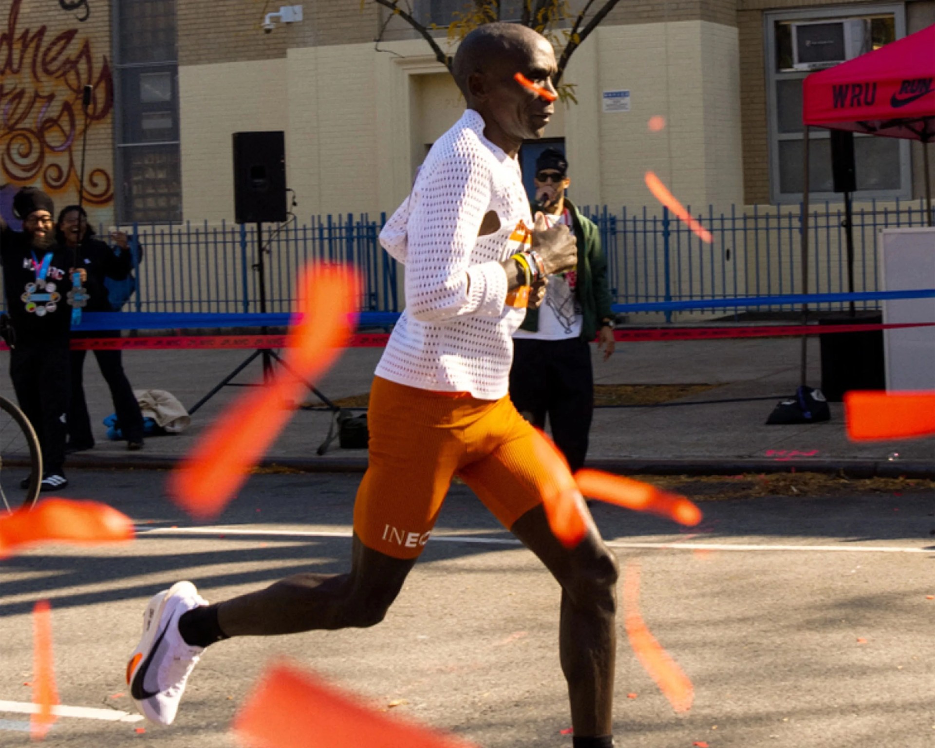 Runner in white perforated long-sleeve shirt and orange shorts crossing a finish line with orange ribbons.