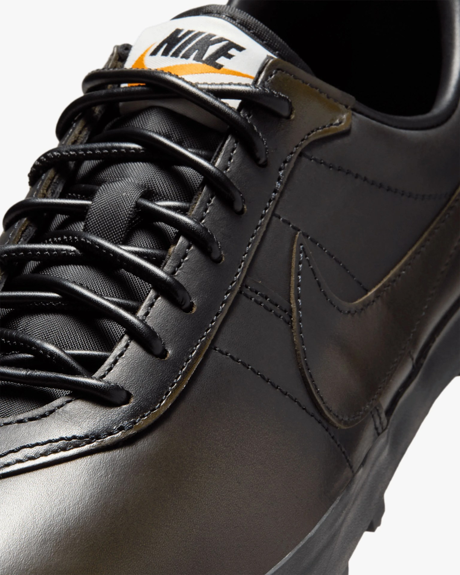 Close-up of black Nike leather sneaker with black laces and white tongue label featuring black and orange Nike logo.