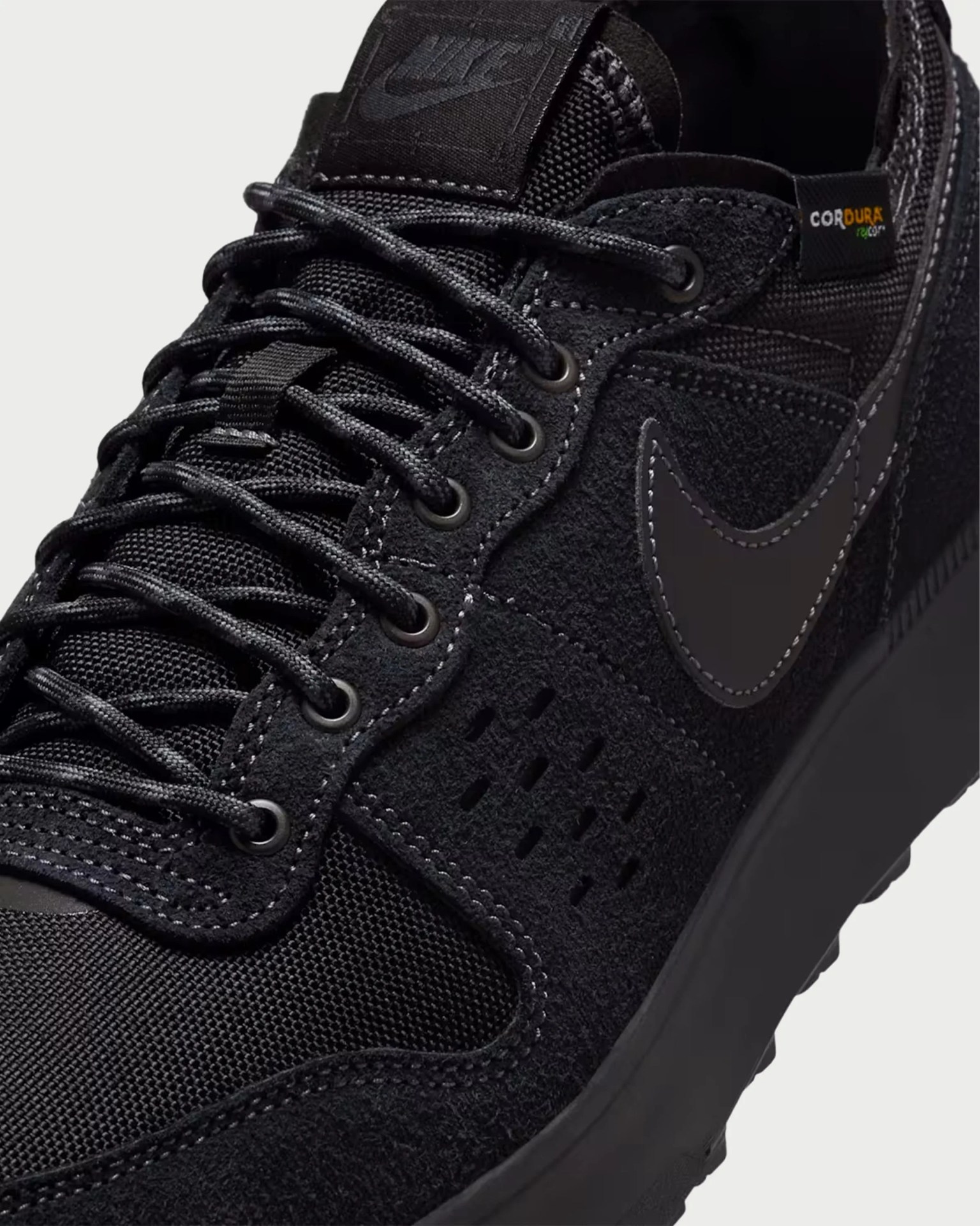 Close-up of a black Nike sneaker with suede and mesh materials, black laces, and a Cordura fabric tag.
