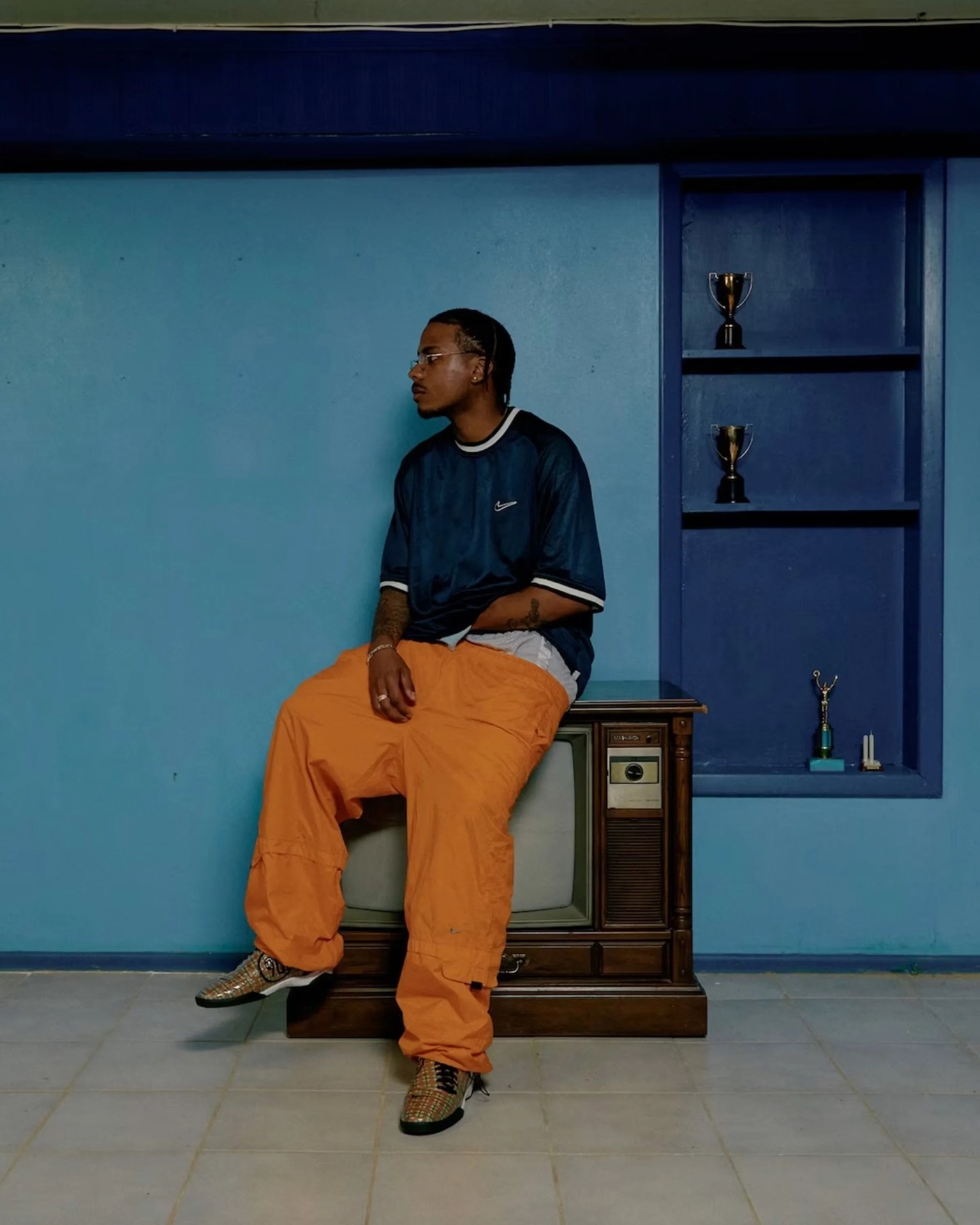 Person wearing orange pants and a dark blue Nike shirt sitting on a vintage wooden TV with blue wall and trophy shelf behind.