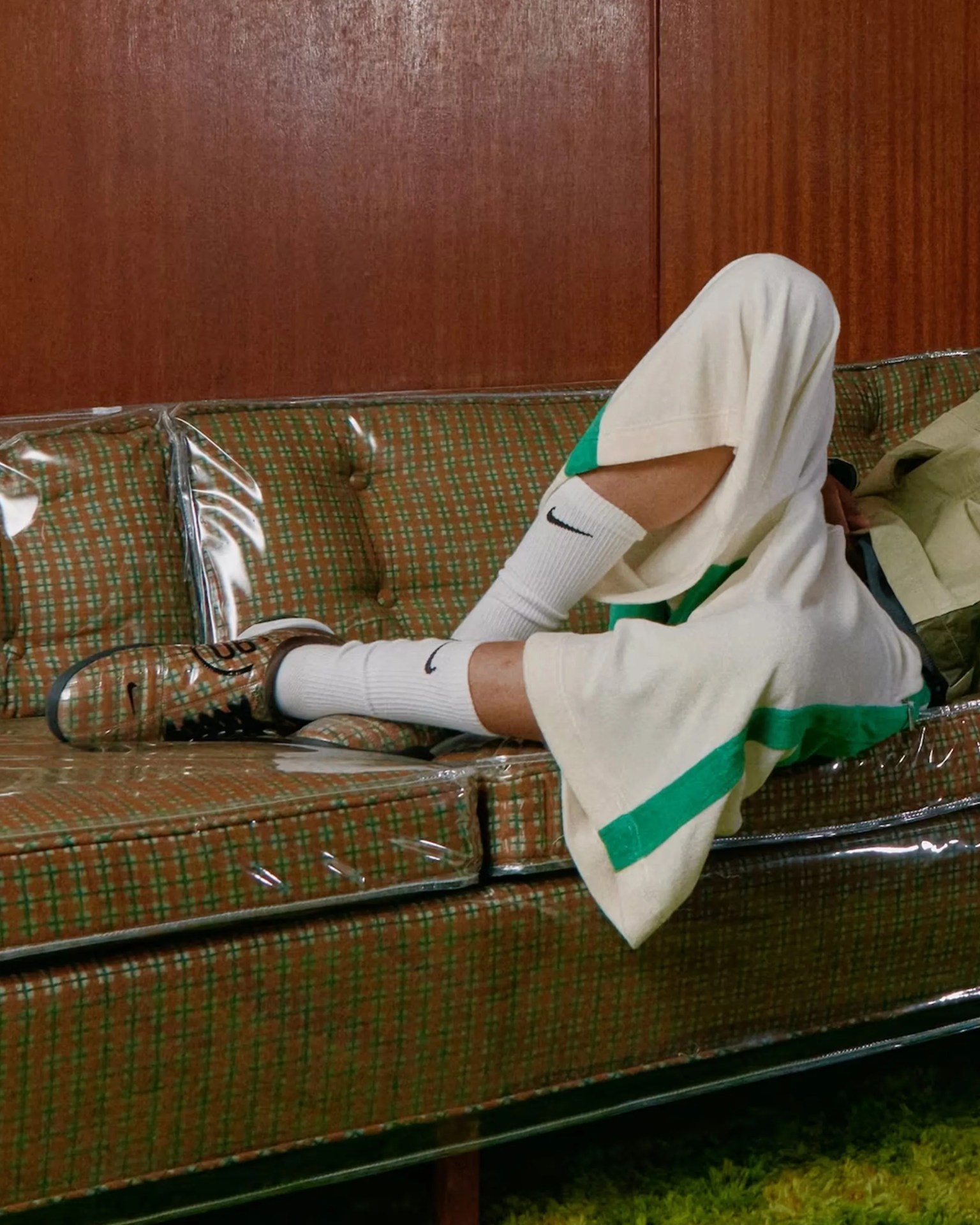 Person wearing cream shorts with green stripes, white Nike socks, and transparent Nike sneakers sitting on a plastic-covered patterned couch.