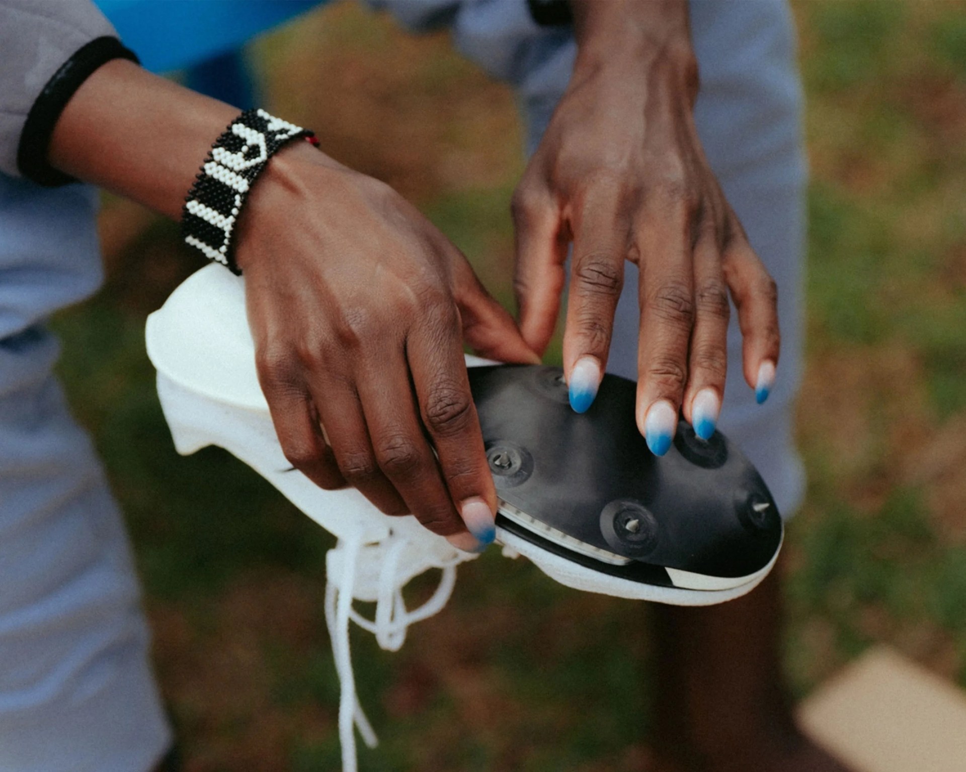 Hands holding a white cleat shoe with black sole and blue-tipped nails, one wrist wearing a black and white beaded bracelet.