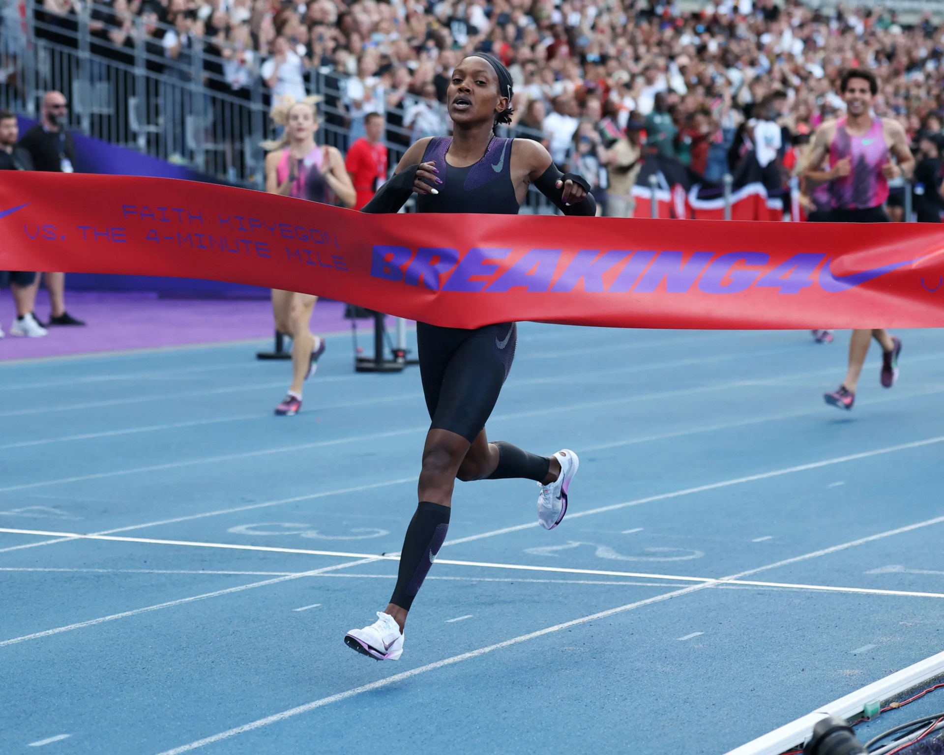 Runner in black Nike athletic wear breaking the red finish line tape on a blue track with blurred crowd in background.