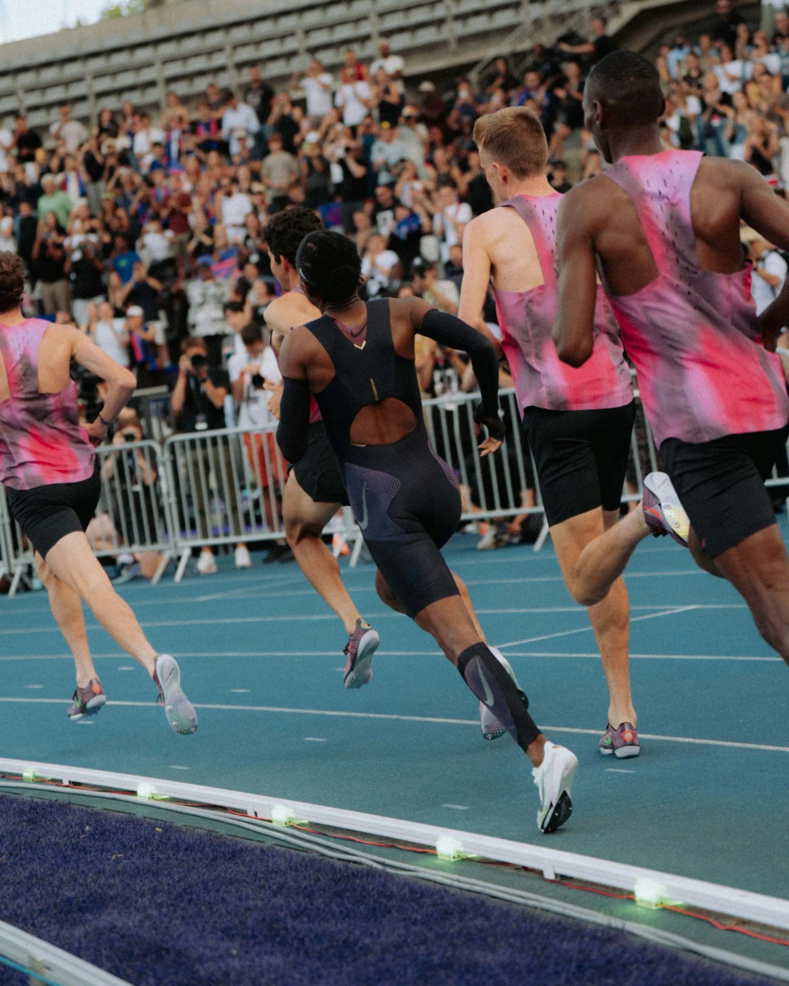 Group of male runners in black and pink athletic gear sprinting on a blue track with a crowd in the background.