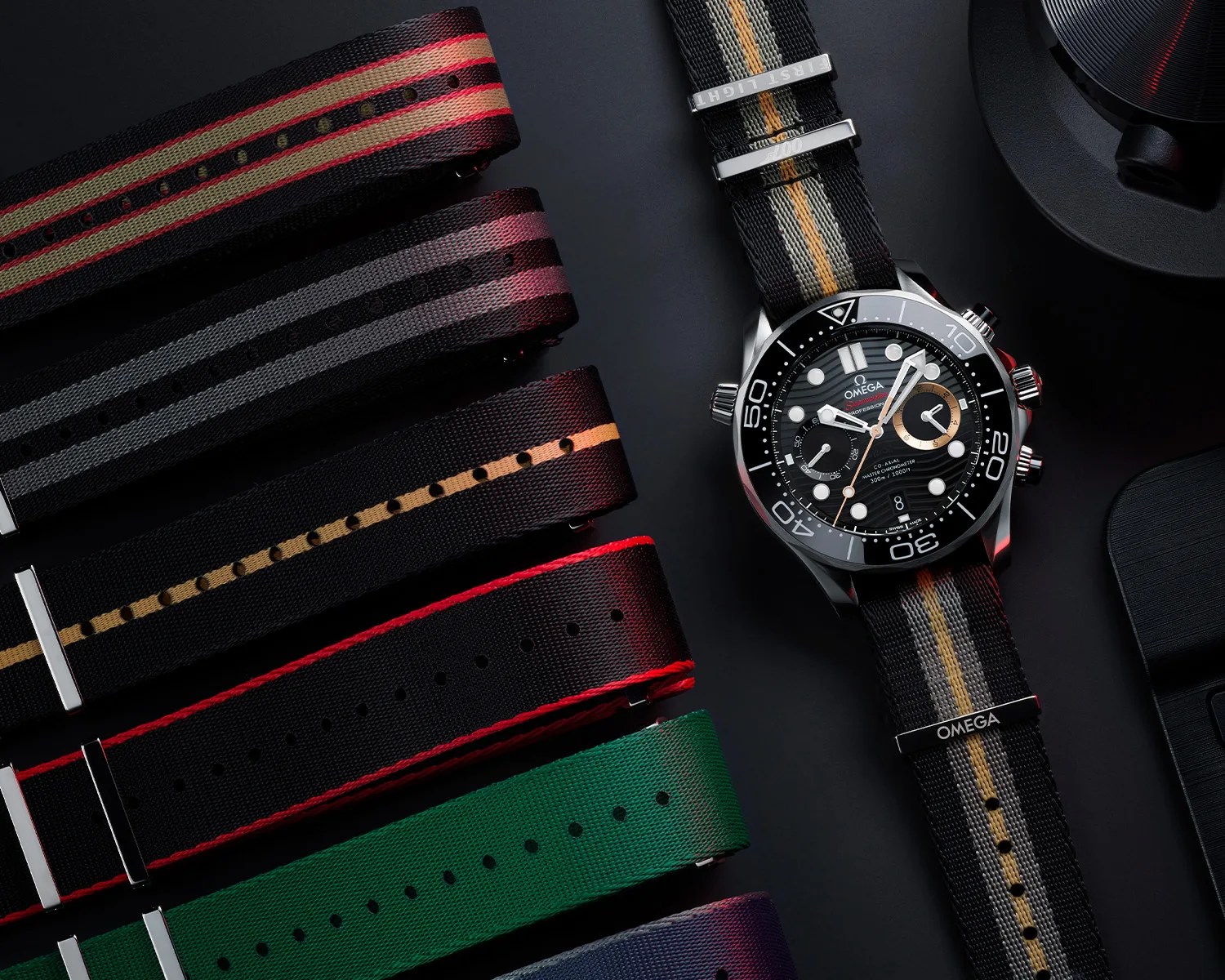 Omega Seamaster watch with black dial and striped black, gray, and gold fabric strap next to five additional striped fabric straps in various colors.