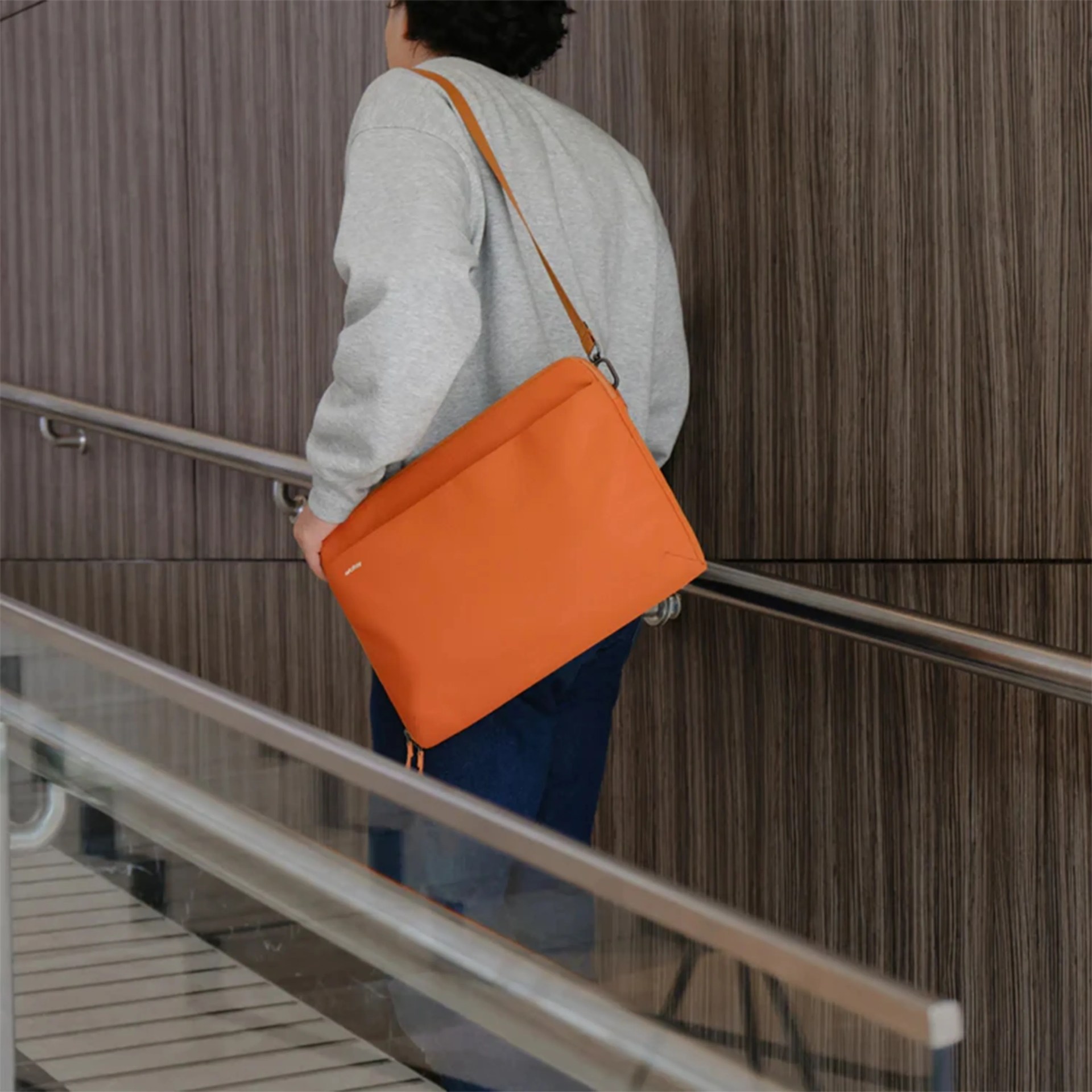 Person wearing a gray sweater and dark pants carrying an orange shoulder bag with a strap.