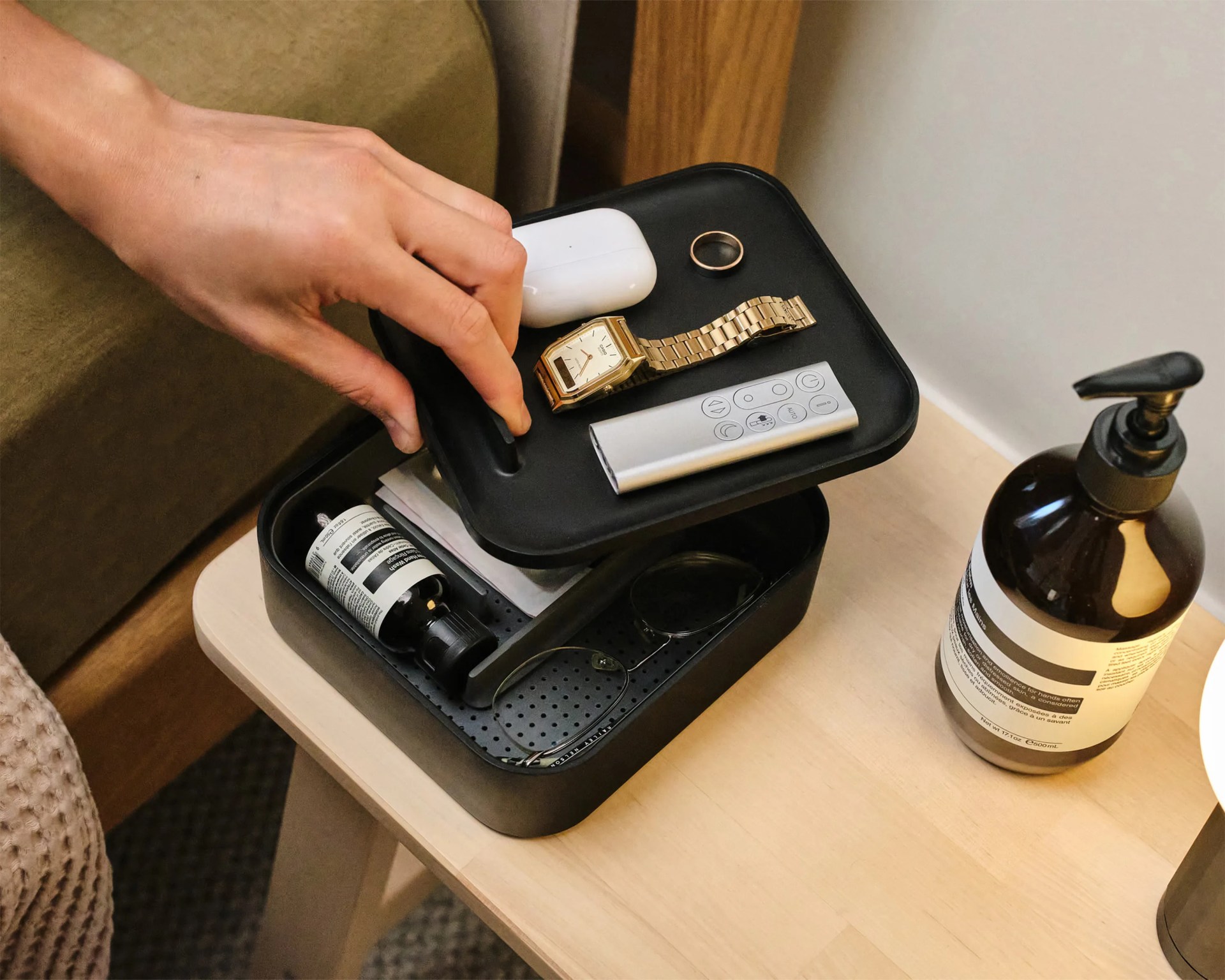 Black organizer tray with a gold wristwatch, white wireless earbuds case, silver remote, black ring, and black sunglasses on a wooden table.