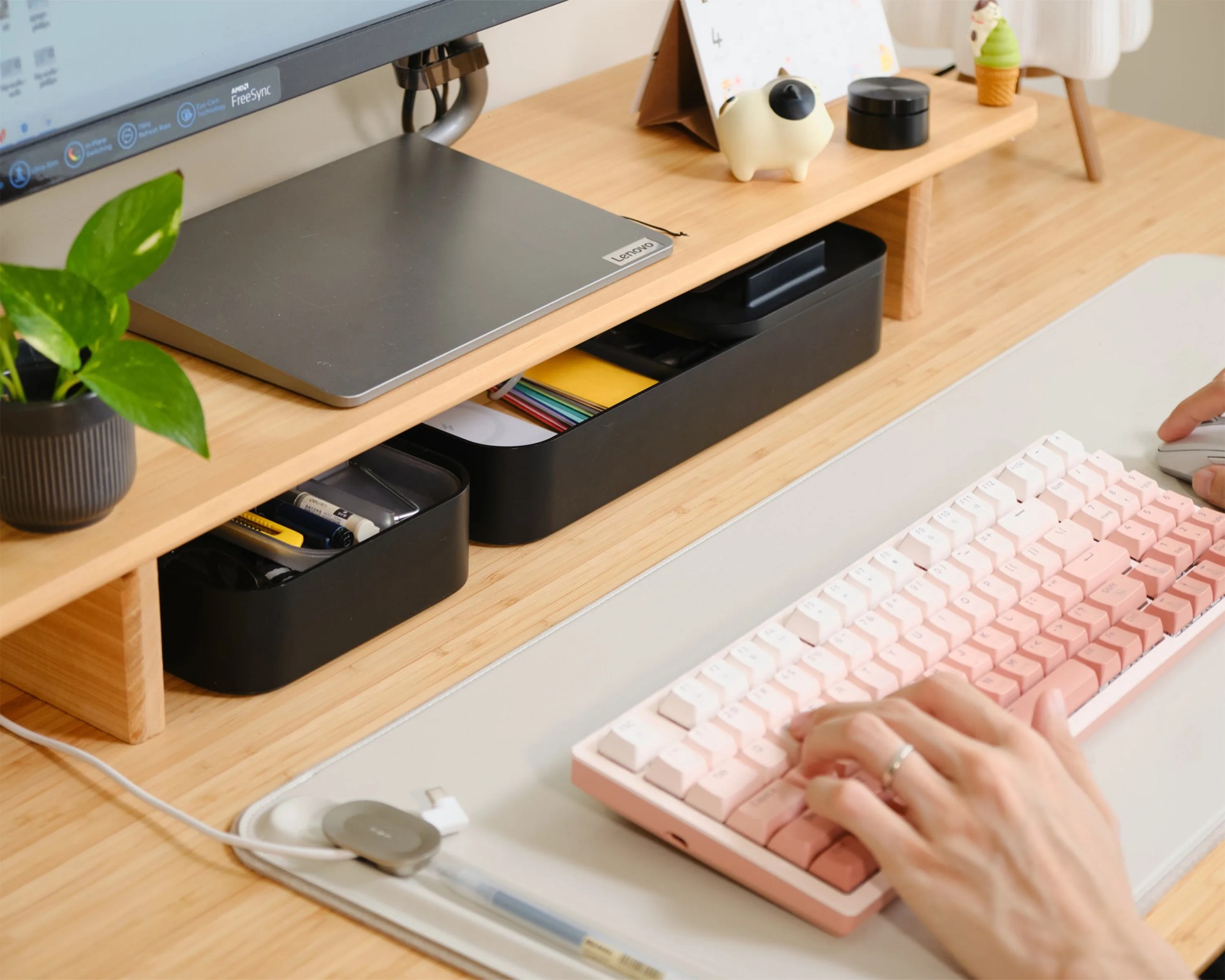 Wooden desk with a Lenovo laptop, black storage trays, a small plant, a pink and white keyboard, and hands typing on the keyboard.