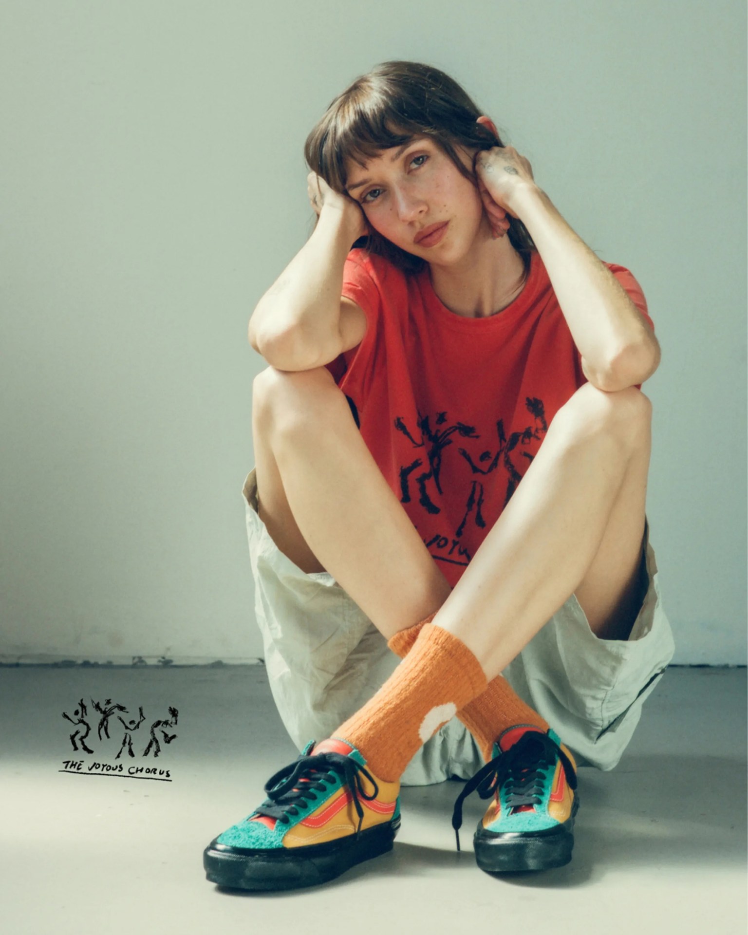 Person sitting on the floor wearing a red graphic t-shirt, light shorts, orange socks, and colorful sneakers.