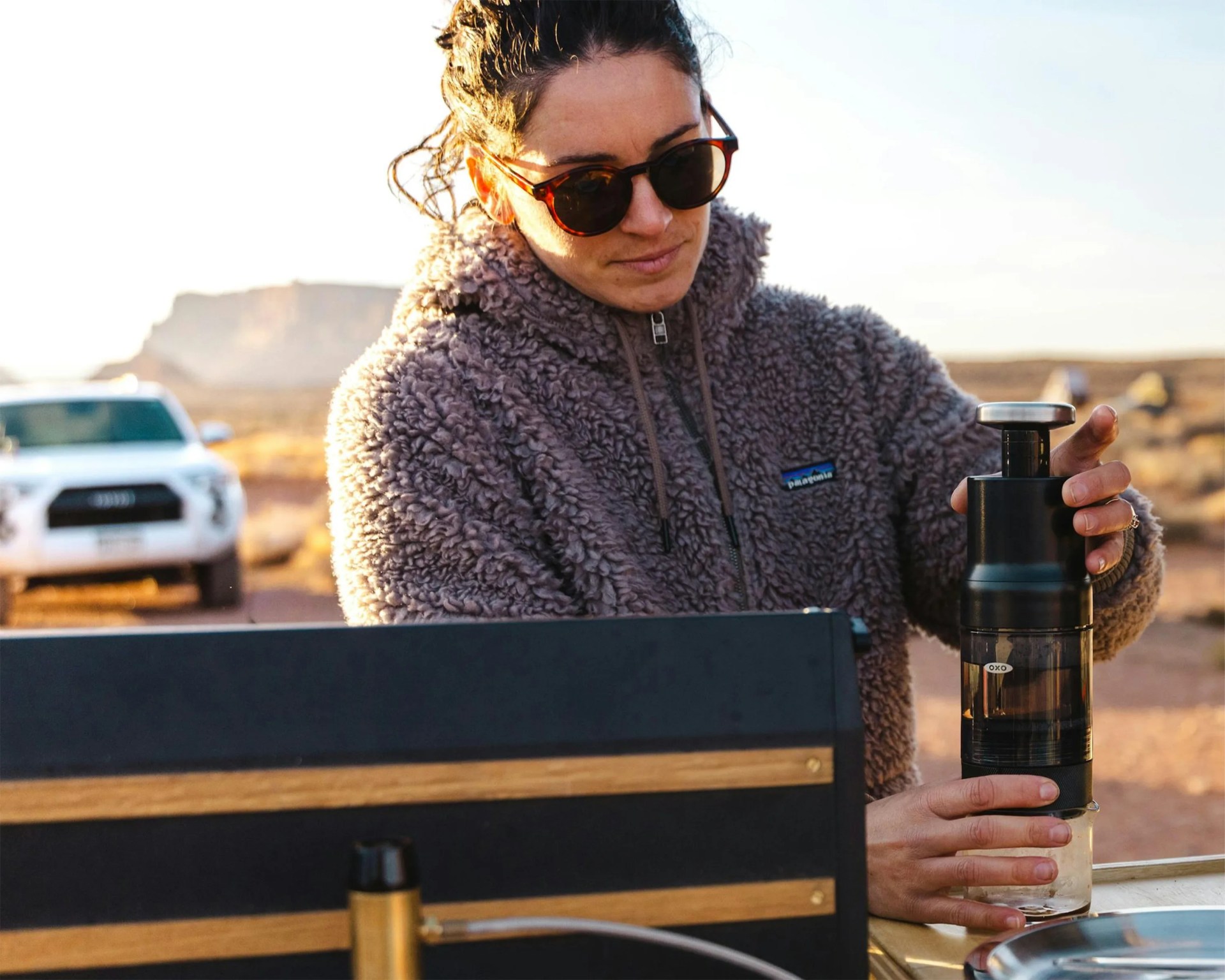 Person wearing sunglasses and a gray Patagonia fleece jacket using a black OXO coffee grinder outdoors with a white vehicle in the background.