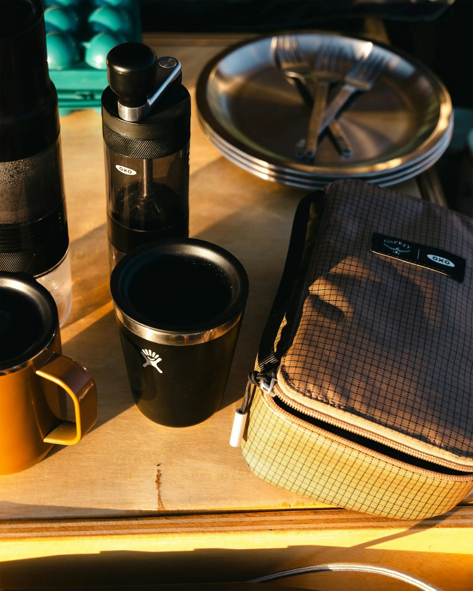 Black OXO manual coffee grinder, black Hydro Flask tumbler, brown metal mug, stack of metal plates with forks, and a brown checkered Osprey and OXO branded bag on a wooden surface.