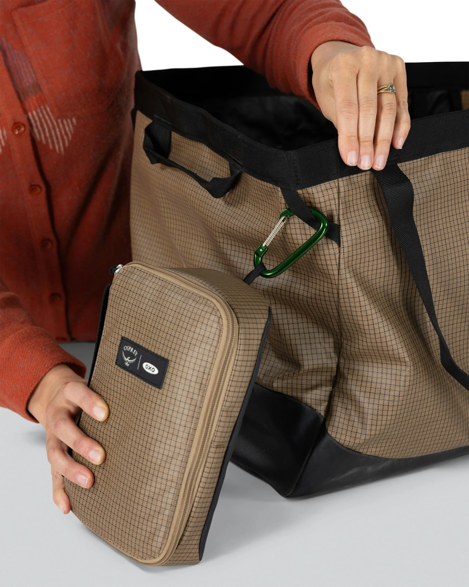 Brown checkered fabric tote bag with black trim and green carabiner, held by a person in an orange shirt.