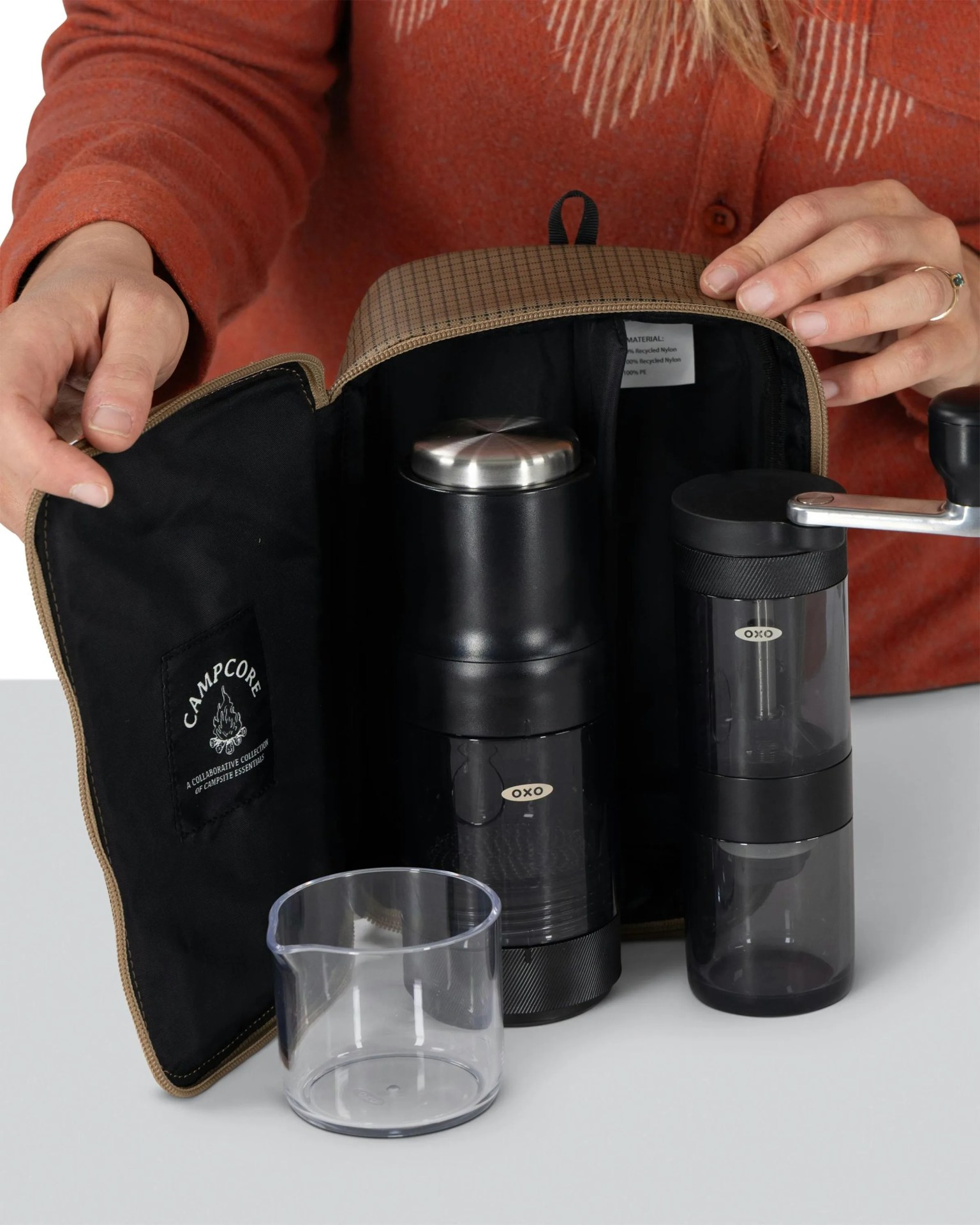 Brown checkered zippered pouch opened to reveal two black OXO coffee grinders and a clear plastic measuring cup on a white surface.