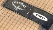 Close-up of a black fabric label with white "Osprey" and "oxo" logos stitched onto tan grid-patterned fabric.