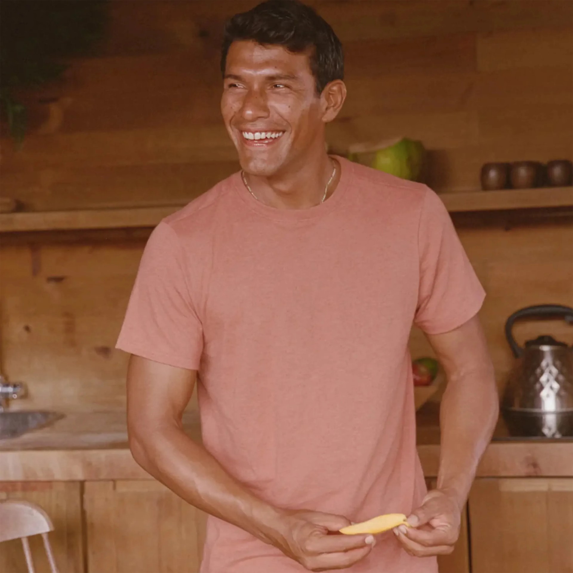 Man in a pink t-shirt smiling and peeling a mango in a wooden kitchen.