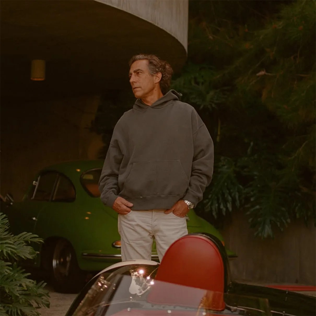 Man wearing a dark gray hoodie and white pants standing near a vintage green car and a red car seat.