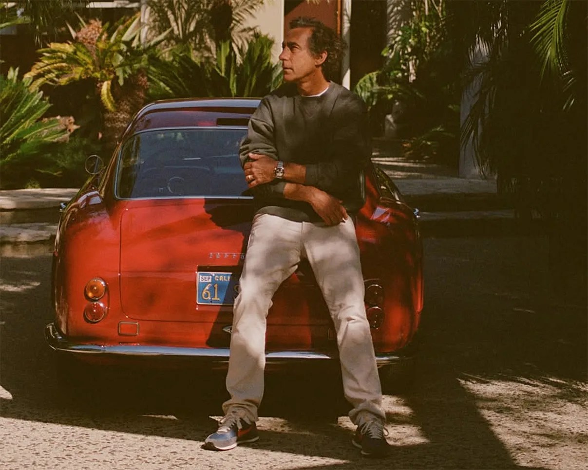 Man wearing a dark sweatshirt and light pants leaning against the rear of a red vintage Ferrari car with a California license plate.