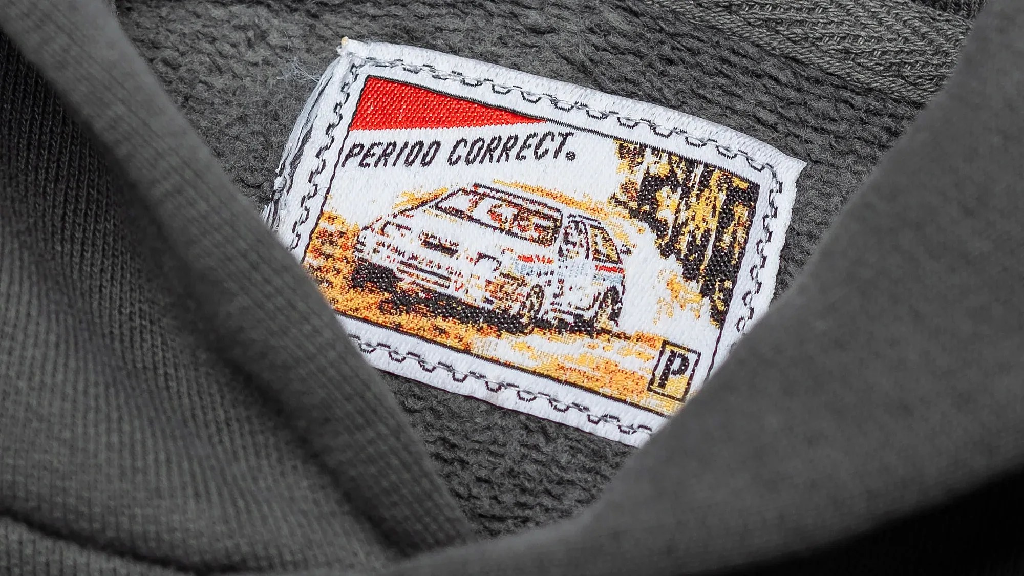 Close-up of a white fabric patch with a rally car illustration and the text "PERIOD CORRECT." sewn onto dark gray textured fabric.