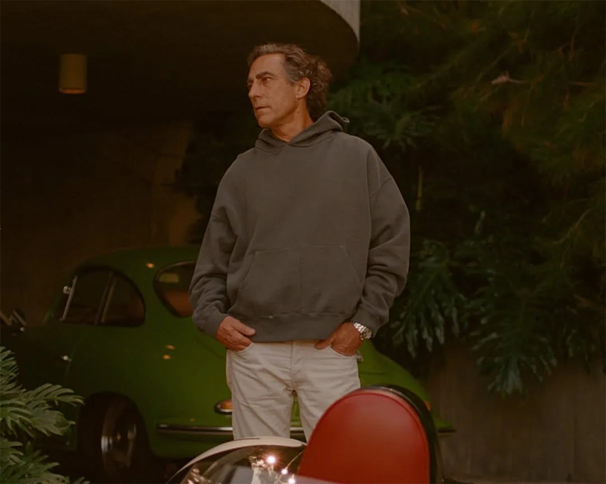 Man wearing a dark gray hoodie and white pants standing in front of a green vintage car.