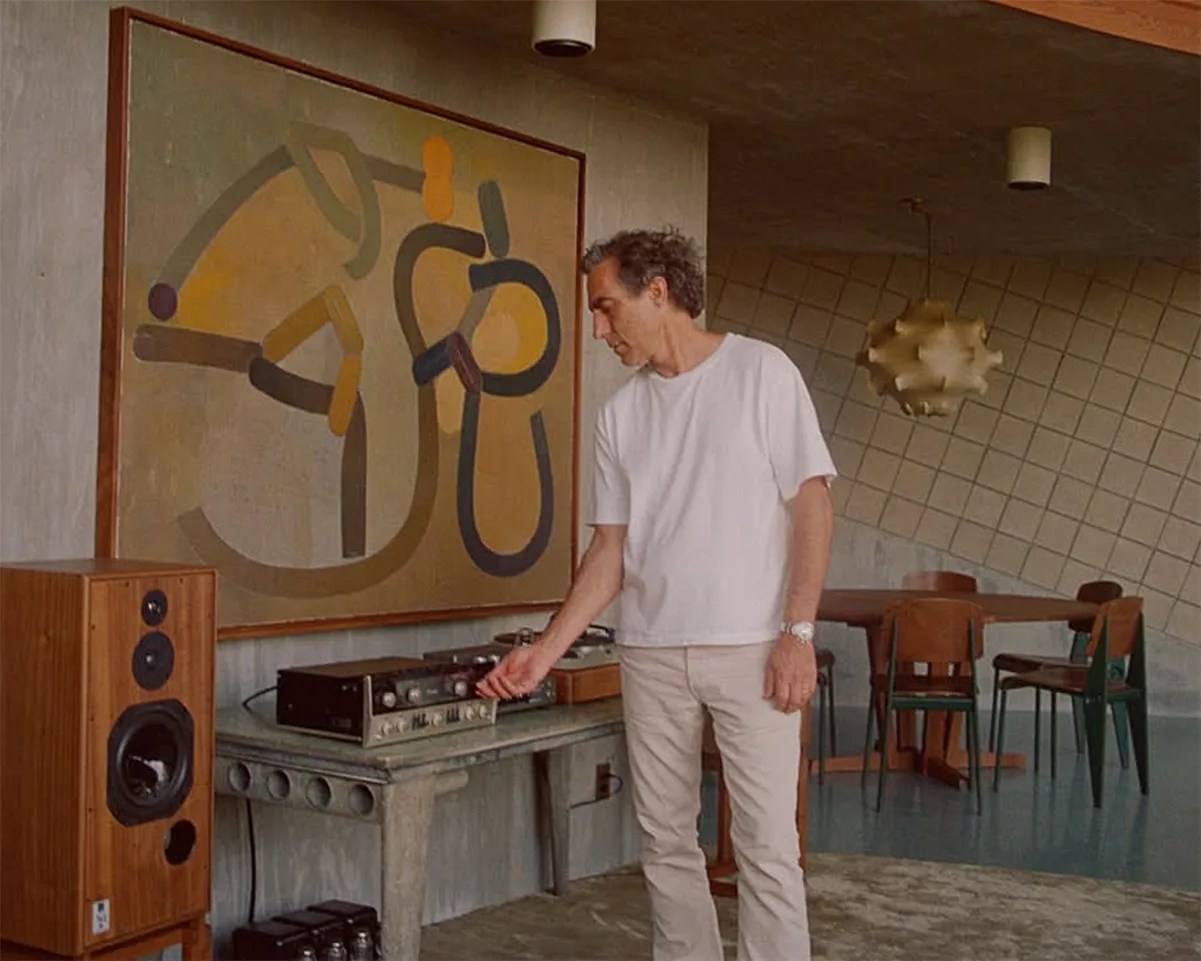 Man in white t-shirt adjusting vintage stereo equipment on a concrete table in a mid-century modern room.