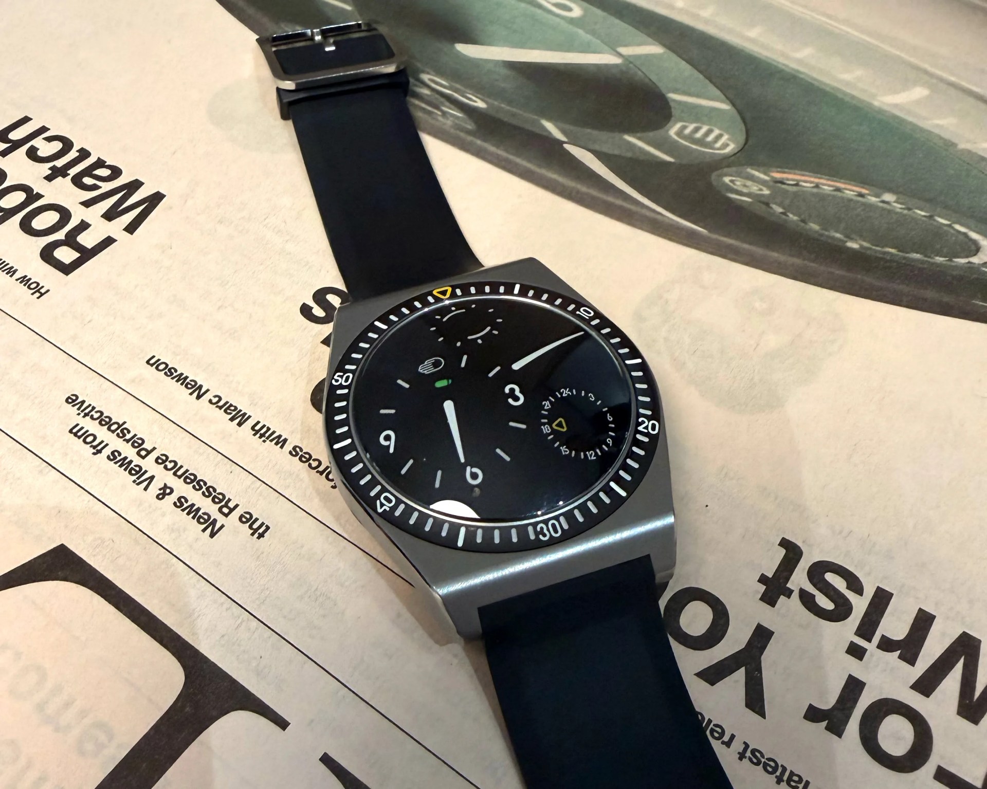 Square silver watch with black dial and black rubber strap resting on a newspaper.