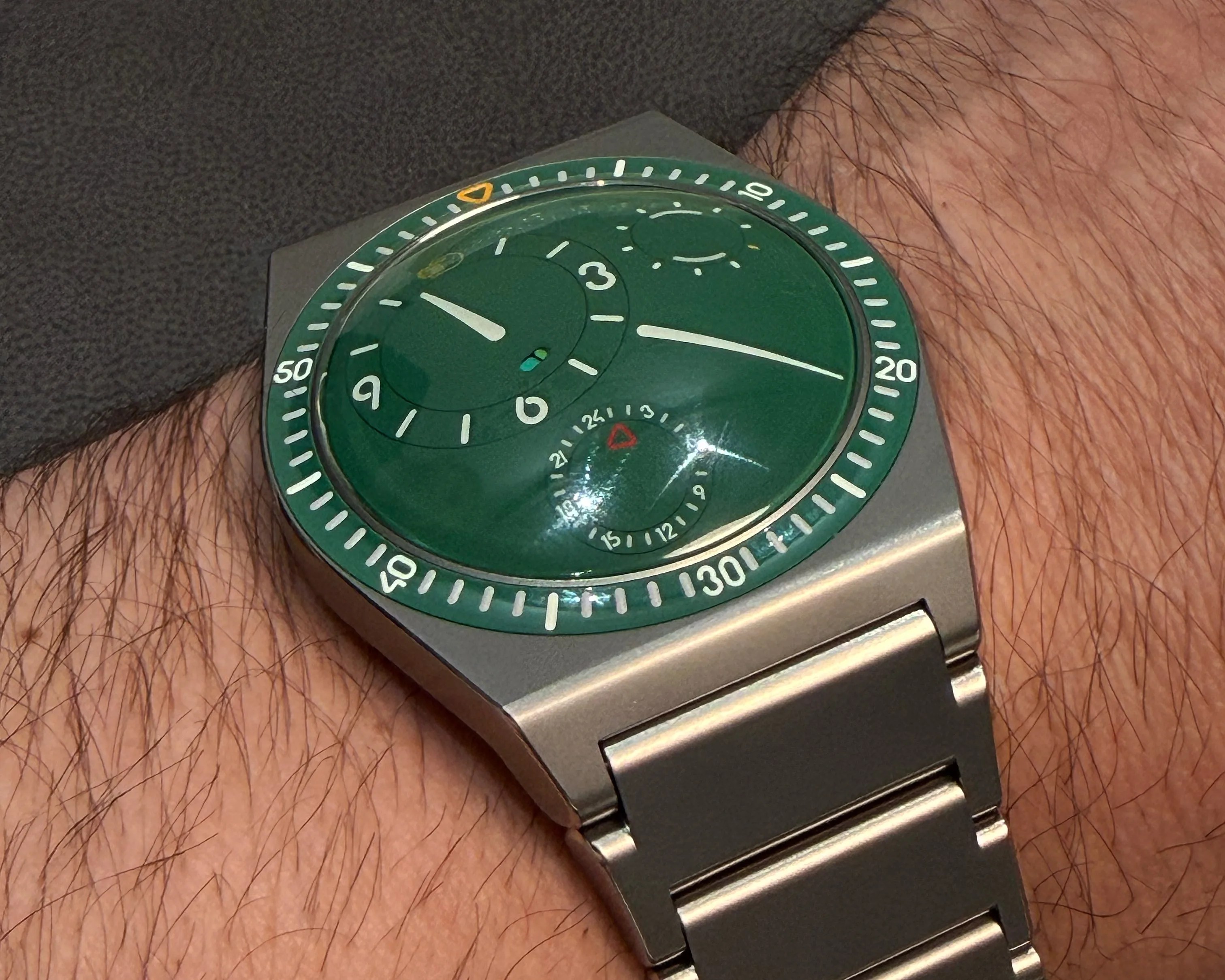 Green dial wristwatch with white markings and a silver metal band on a wrist.