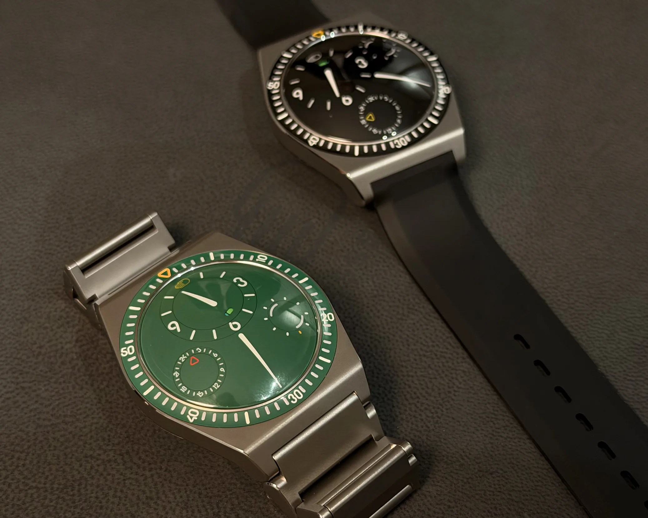 Two wristwatches with round dials, one with a green face and metal bracelet, the other with a black face and black strap, on a dark surface.