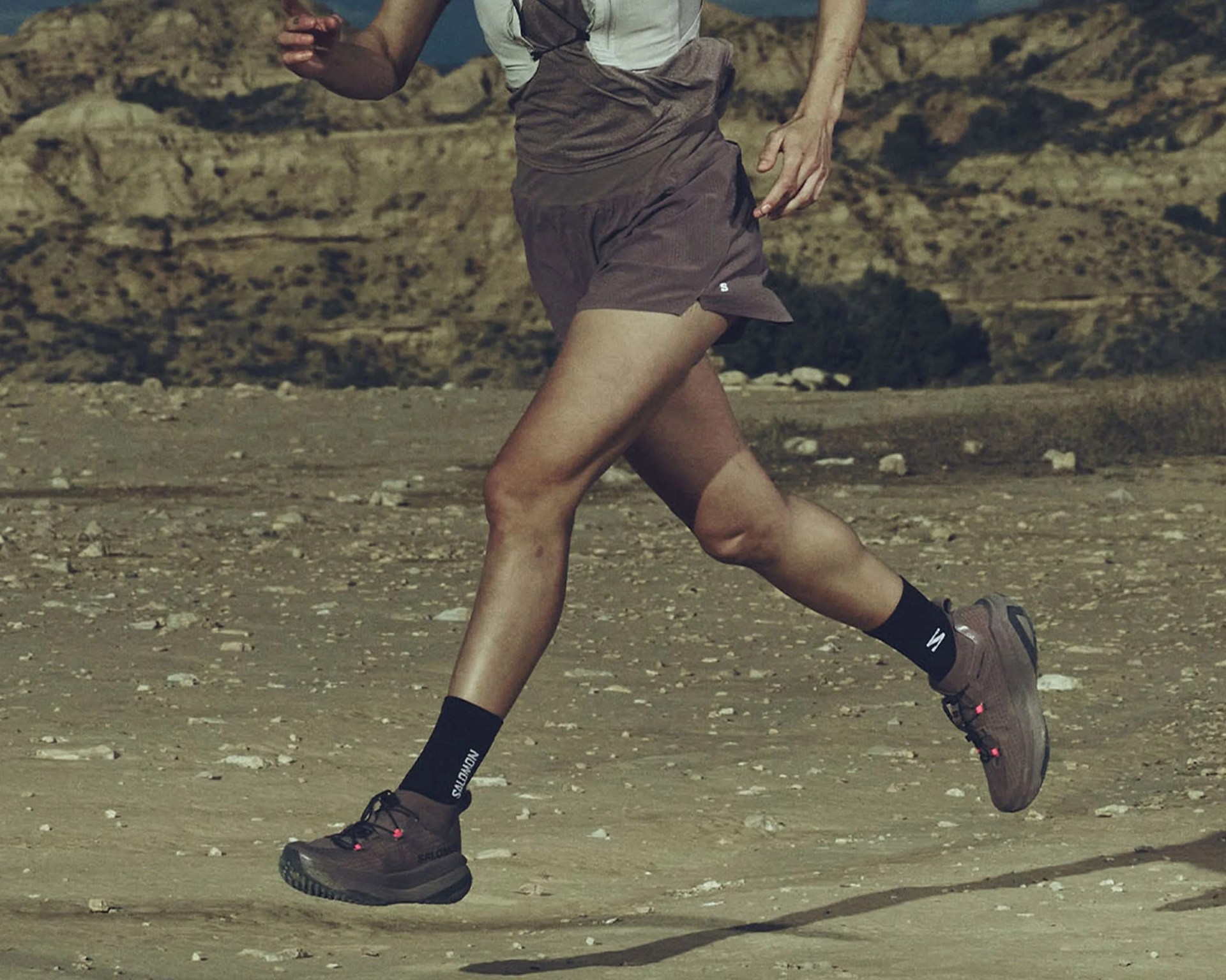 Person running on a rocky trail wearing brown trail running shoes and black socks with "SALOMON" branding.