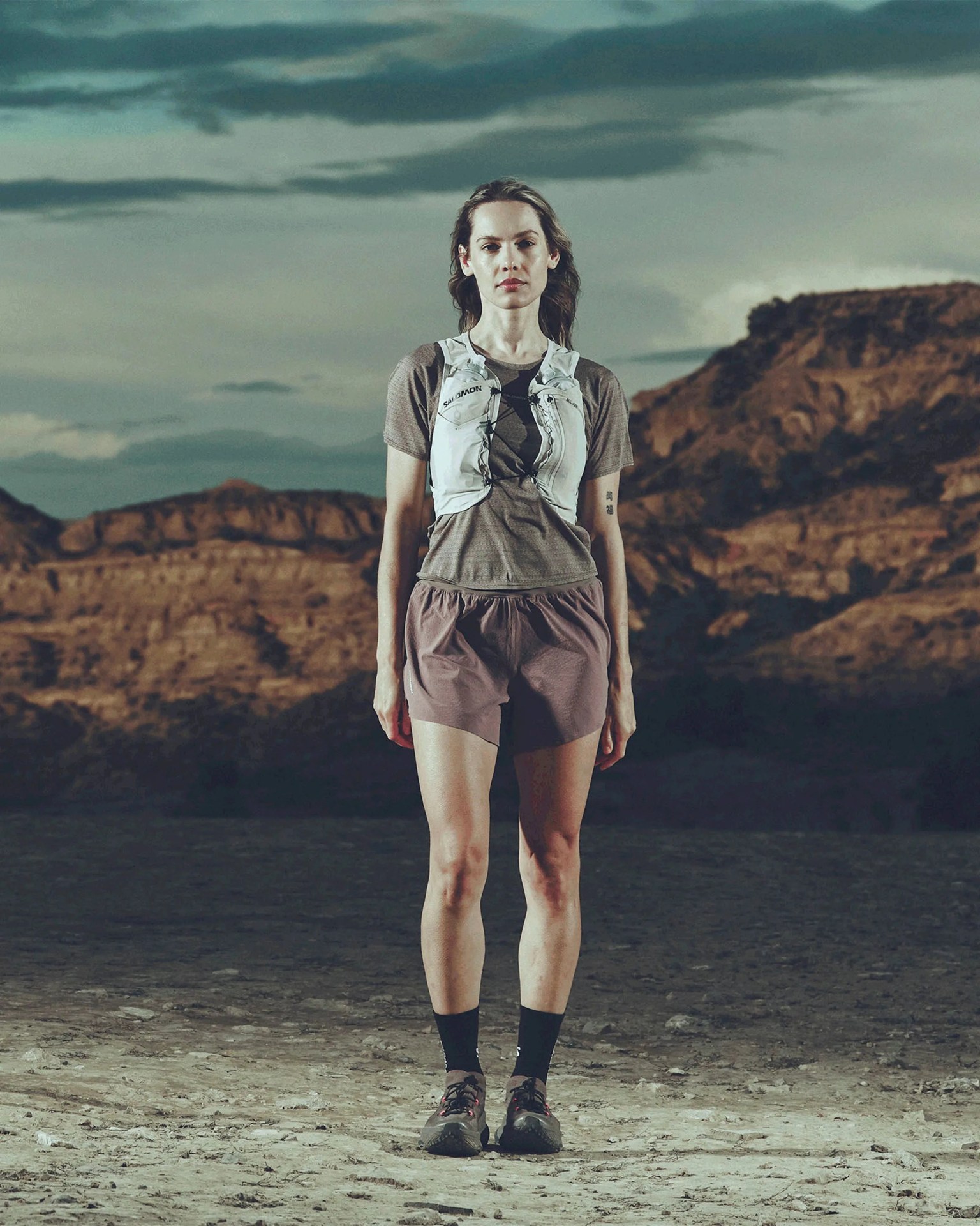 Woman wearing a gray short-sleeve shirt, mauve shorts, black socks, trail running shoes, and a white hydration vest standing on rocky terrain.