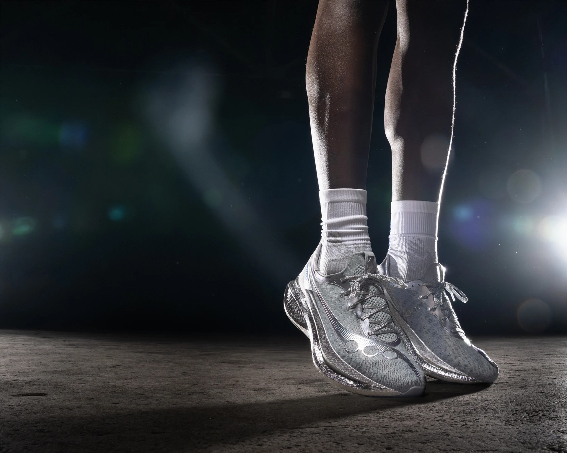 Close-up of legs wearing white socks and silver running shoes on a dark textured surface with bright backlighting.