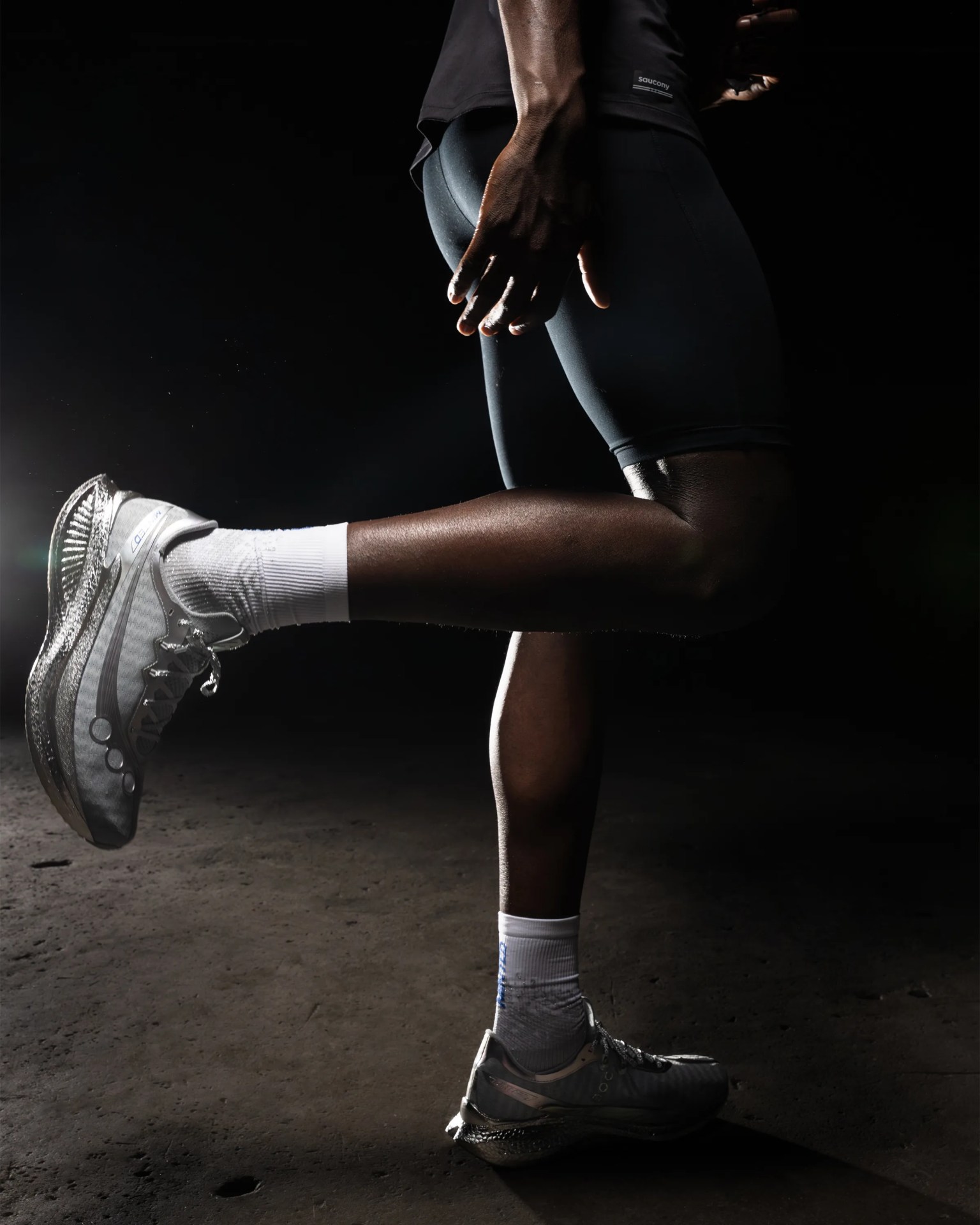 Person wearing black athletic shorts and white running shoes stretching one leg backward on a dark surface.