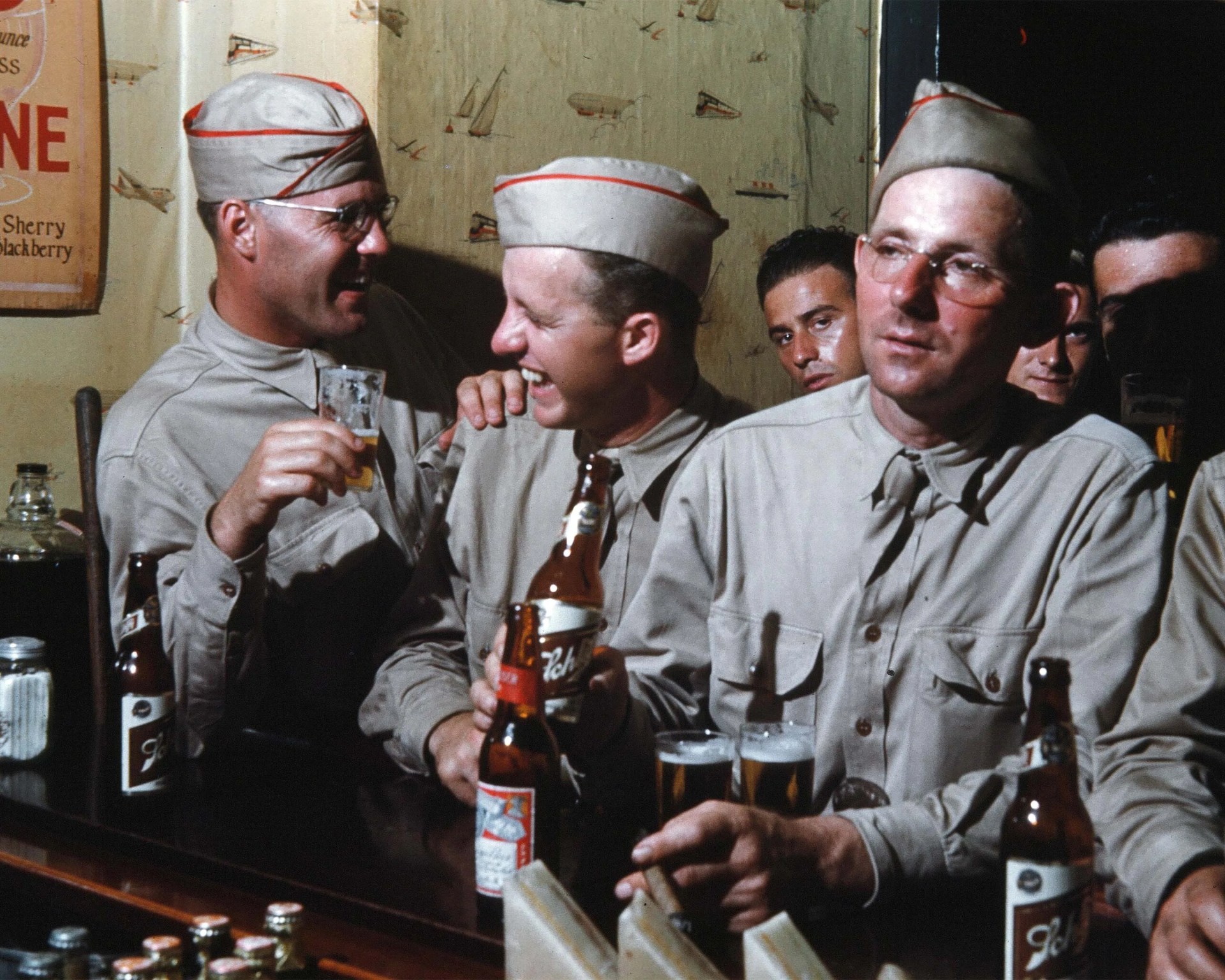 Group of men in beige military uniforms and garrison caps sitting at a bar with beer bottles and glasses.