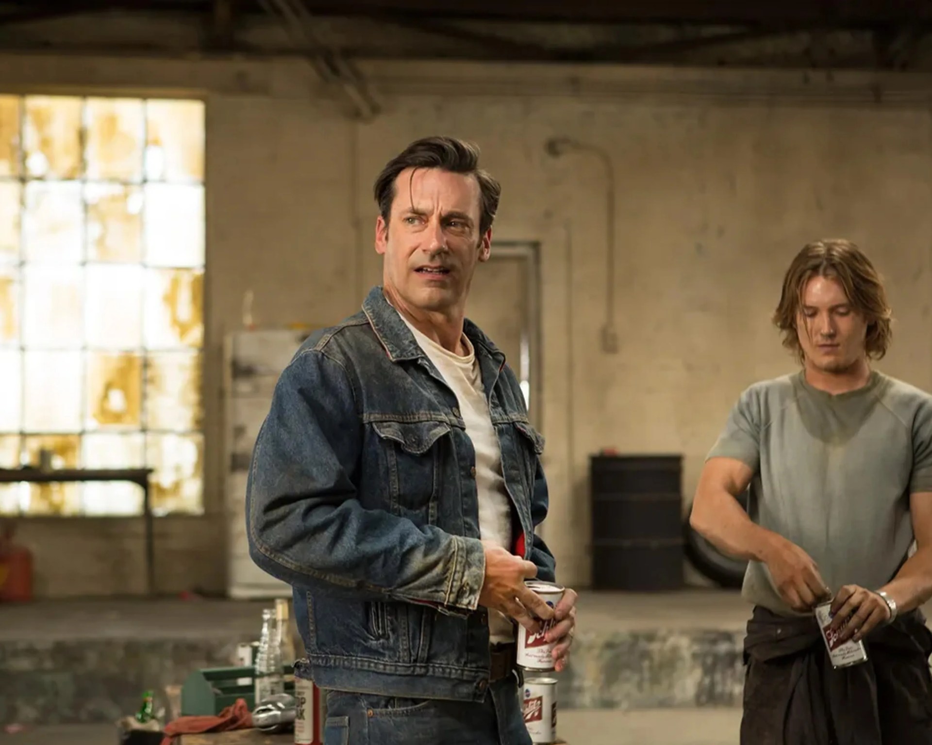 Two men in a rustic industrial room, one wearing a denim jacket and holding a can of Budweiser beer.